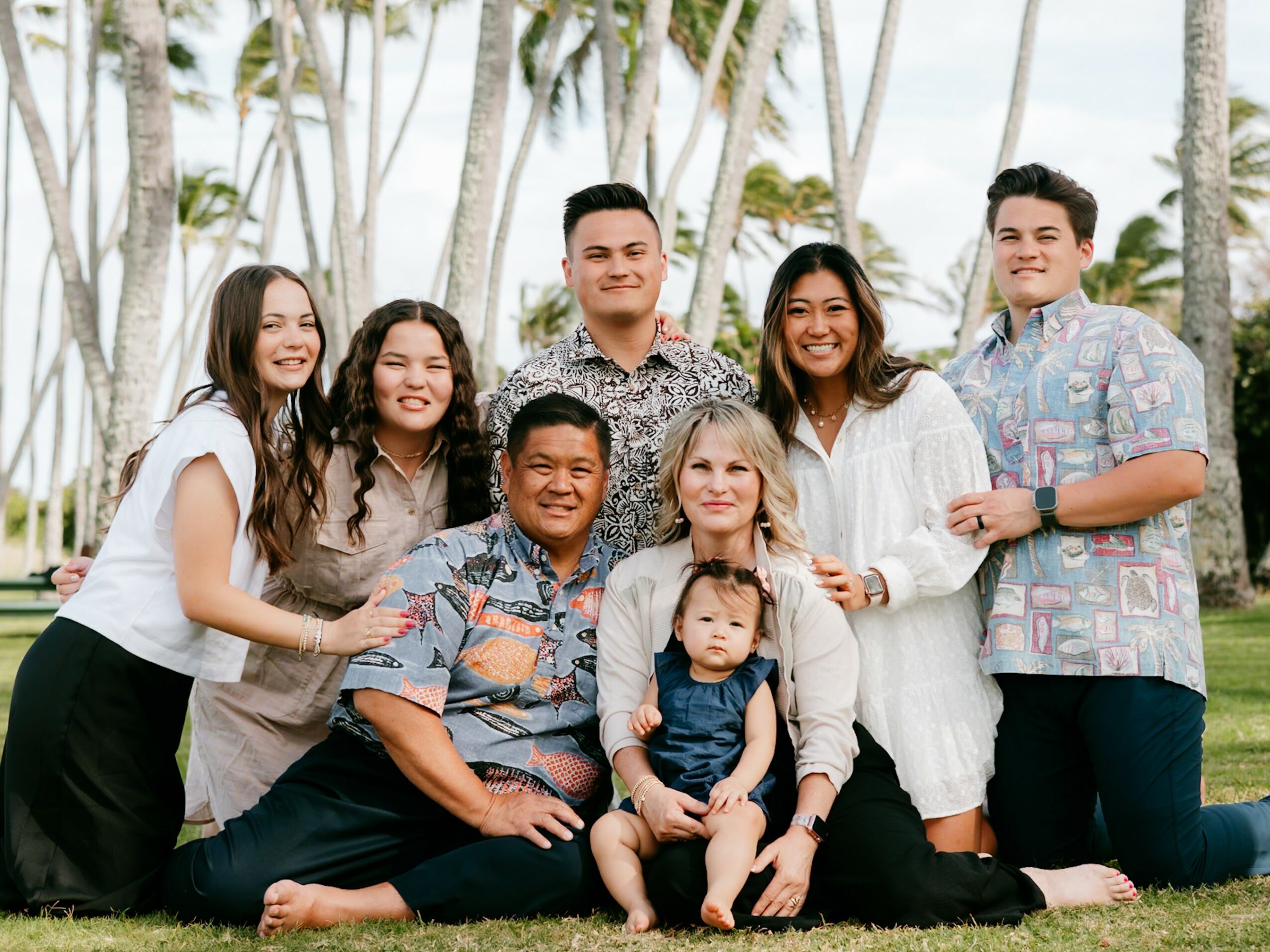 Group of seven people, including children, sitting outdoors with palm trees in background, smiling and posing for photo.