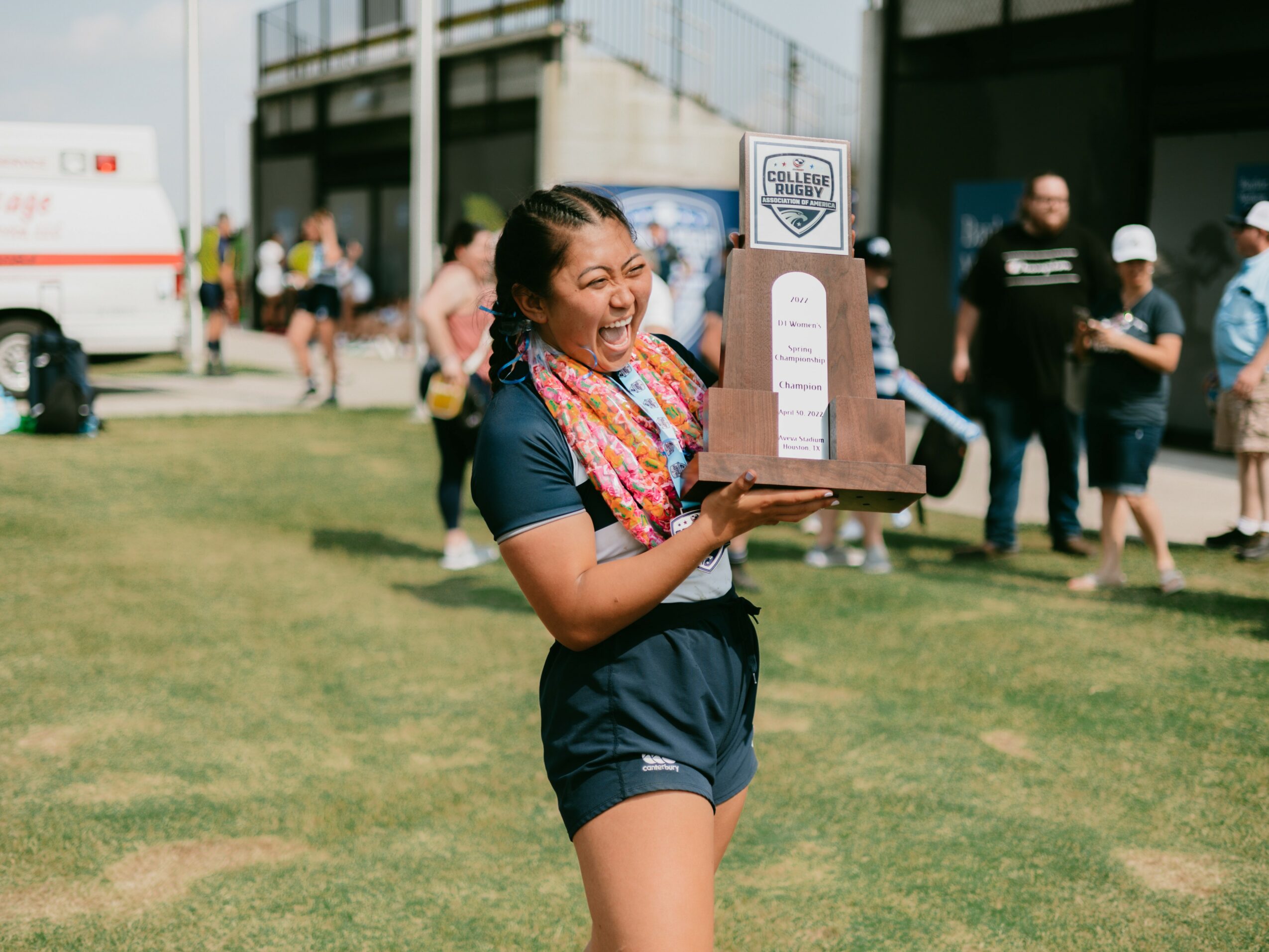 Young woman holding a trophy, smiling, outdoors with people and buildings in background.