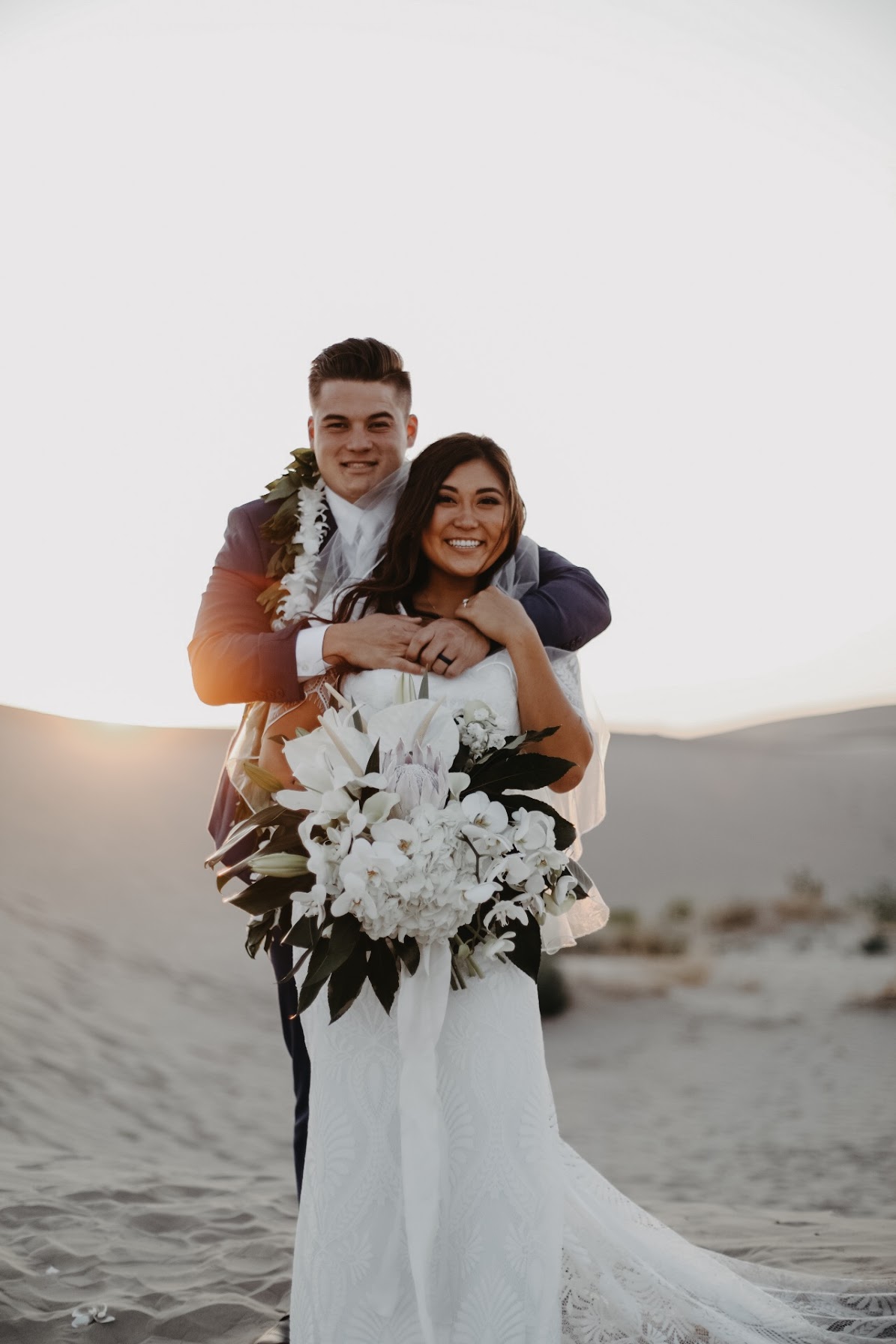 A couple on a beach, with the woman holding a large bouquet of white flowers, smiling at the camera.