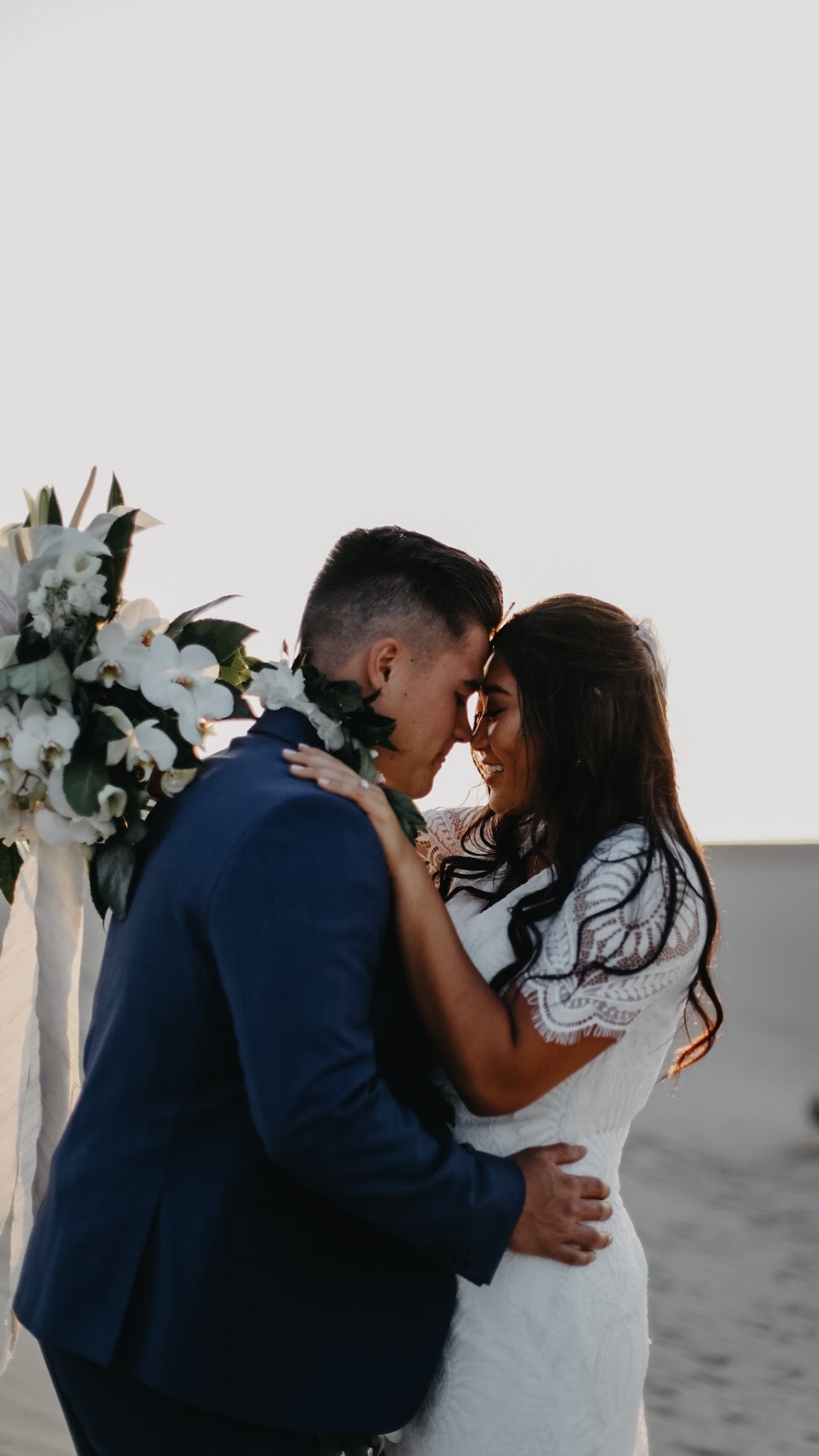 A couple stands close together outdoors, touching foreheads with eyes closed, surrounded by flowers, during sunset or sunrise.
