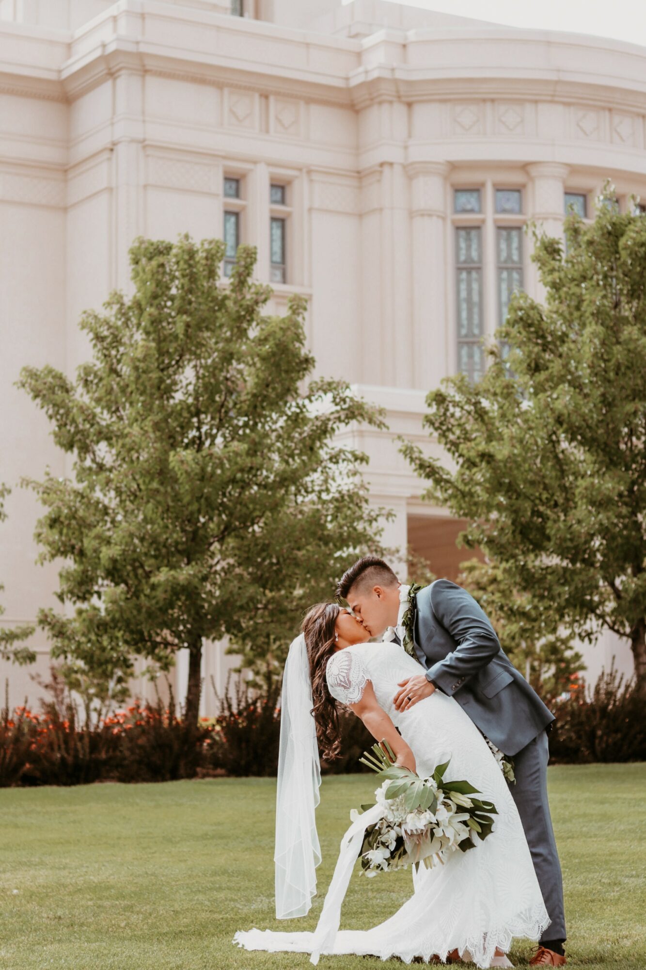 A bride and groom kiss outdoors in front of a historic building with trees, the bride holding a bouquet.