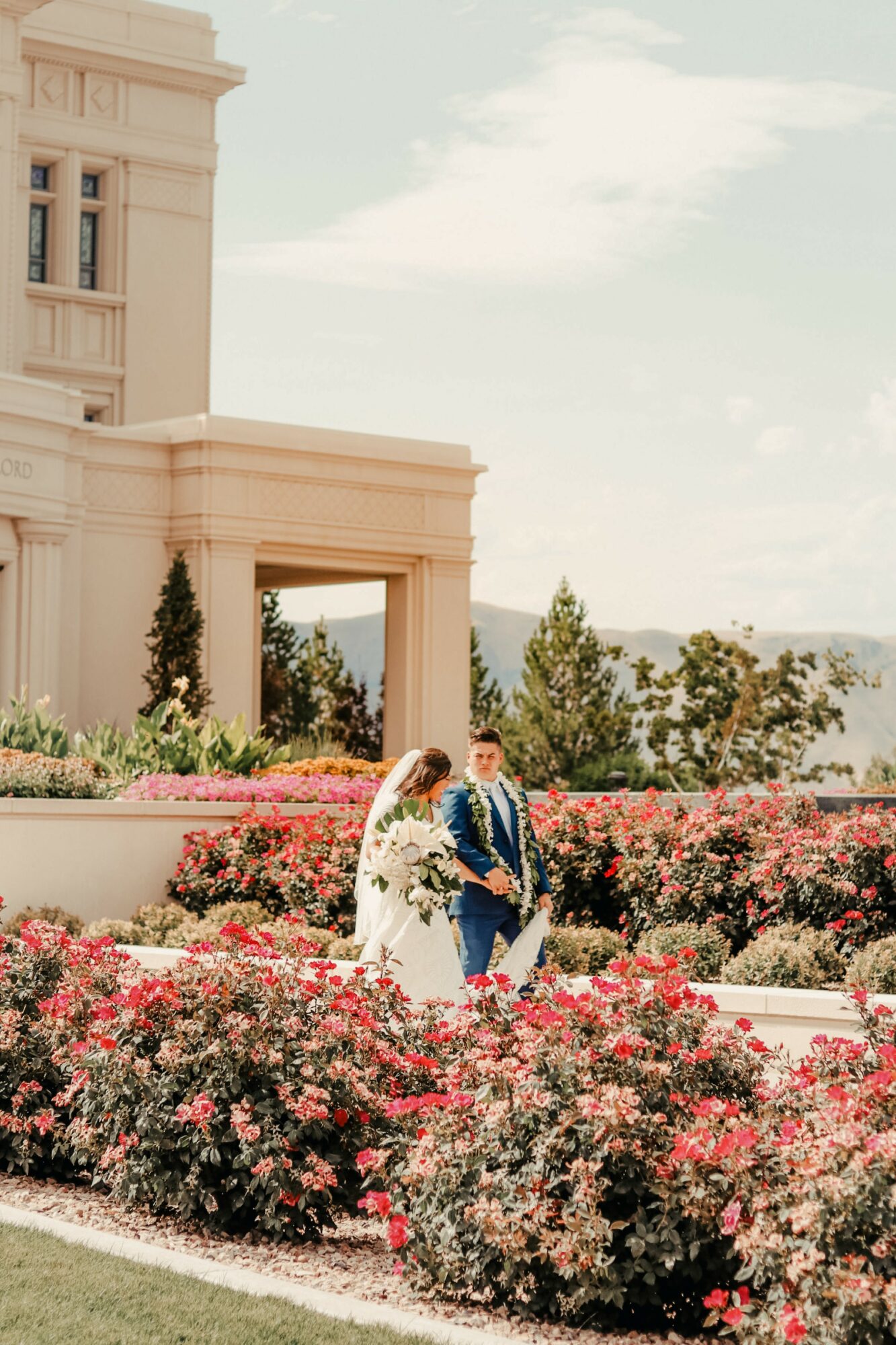 A bride and groom sit on a bench surrounded by pink and red flowers in front of a large building with trees and sky in background.