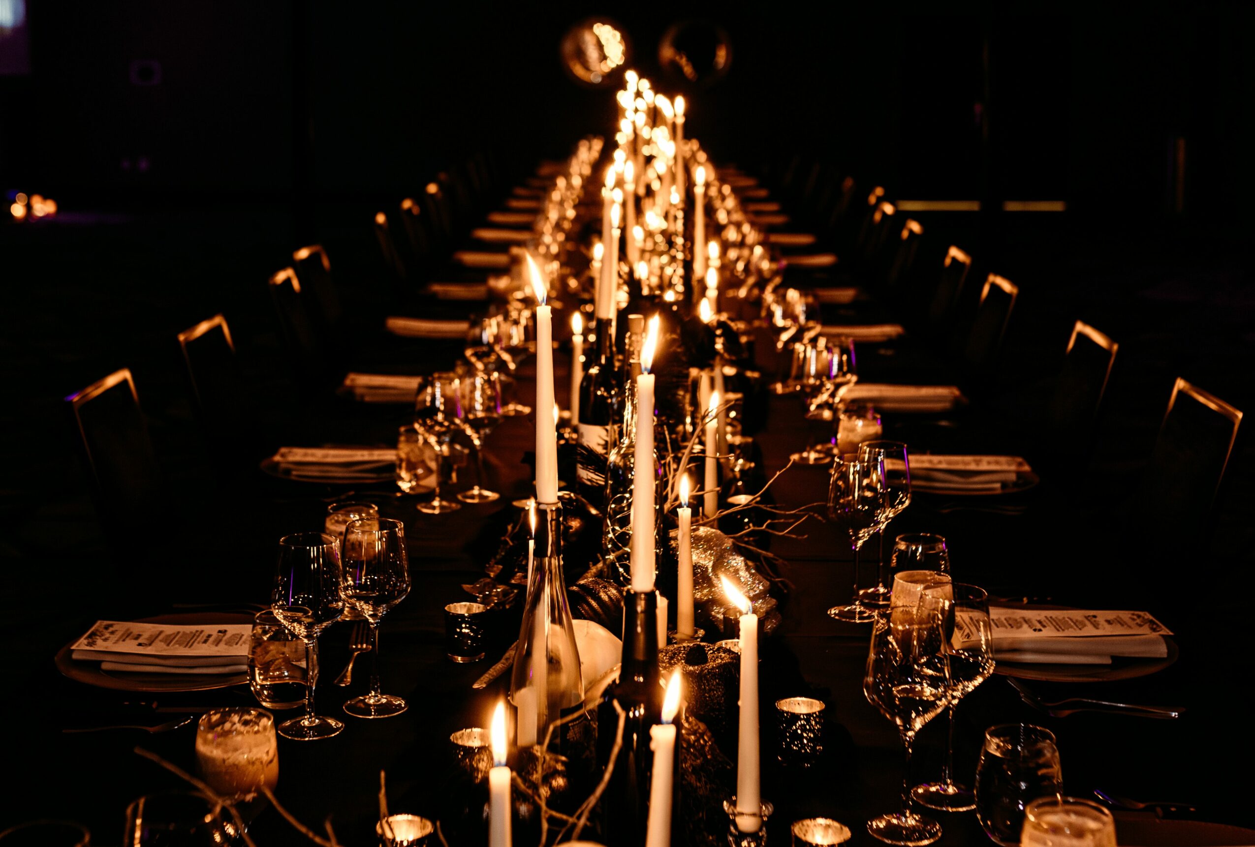 Long dining table decorated with candles and glassware, dimly lit, elegant setting, with chairs on both sides.