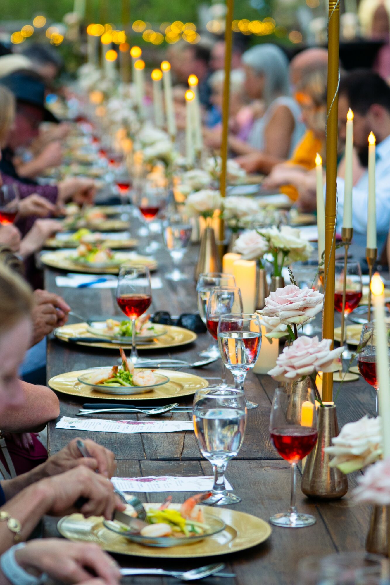 Long outdoor table set with plates, glasses, candles, and people dining, with blurred background of umbrellas and trees.