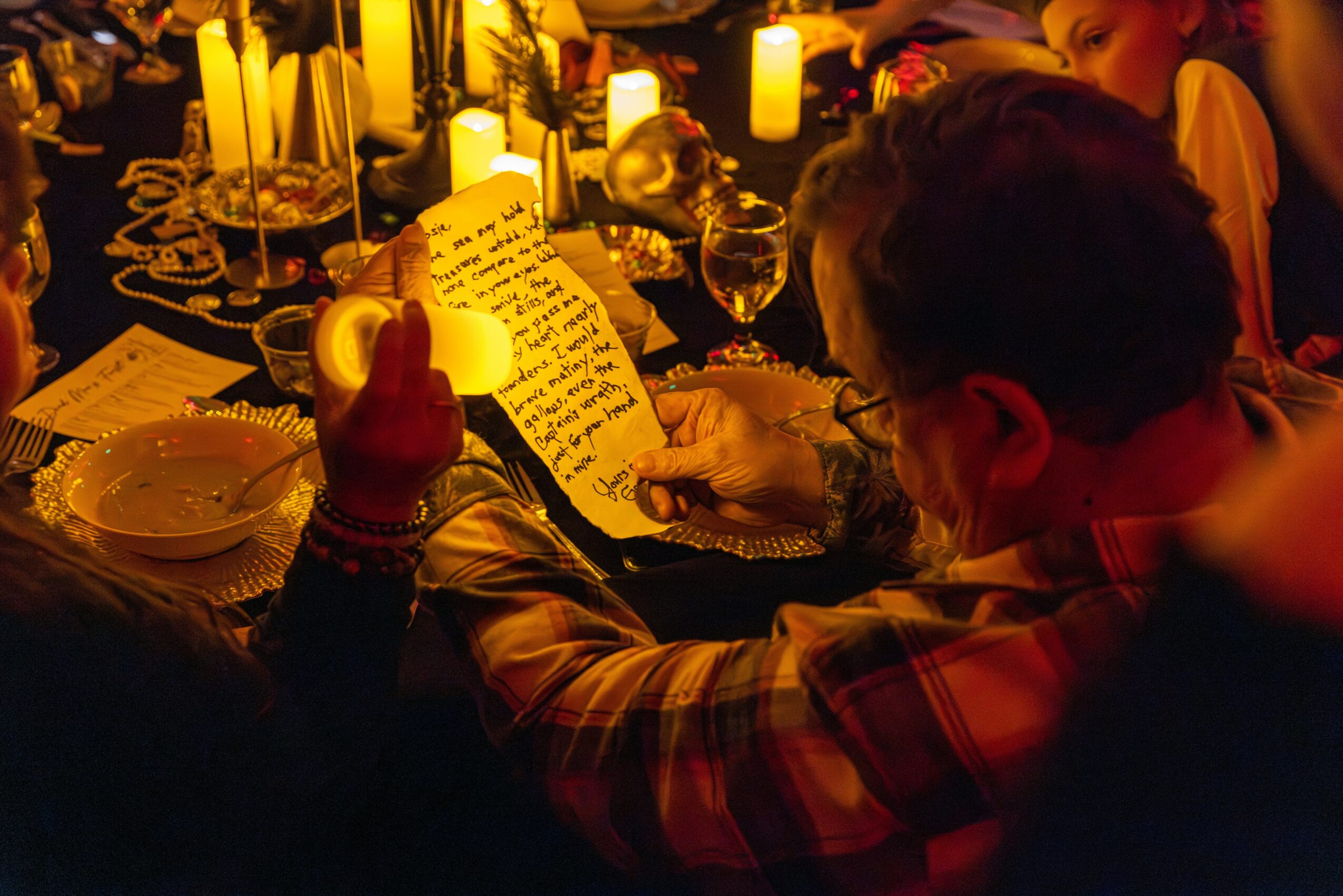 People reading handwritten notes at a candlelit dinner table with plates and glasses, warm lighting, and a cozy atmosphere.