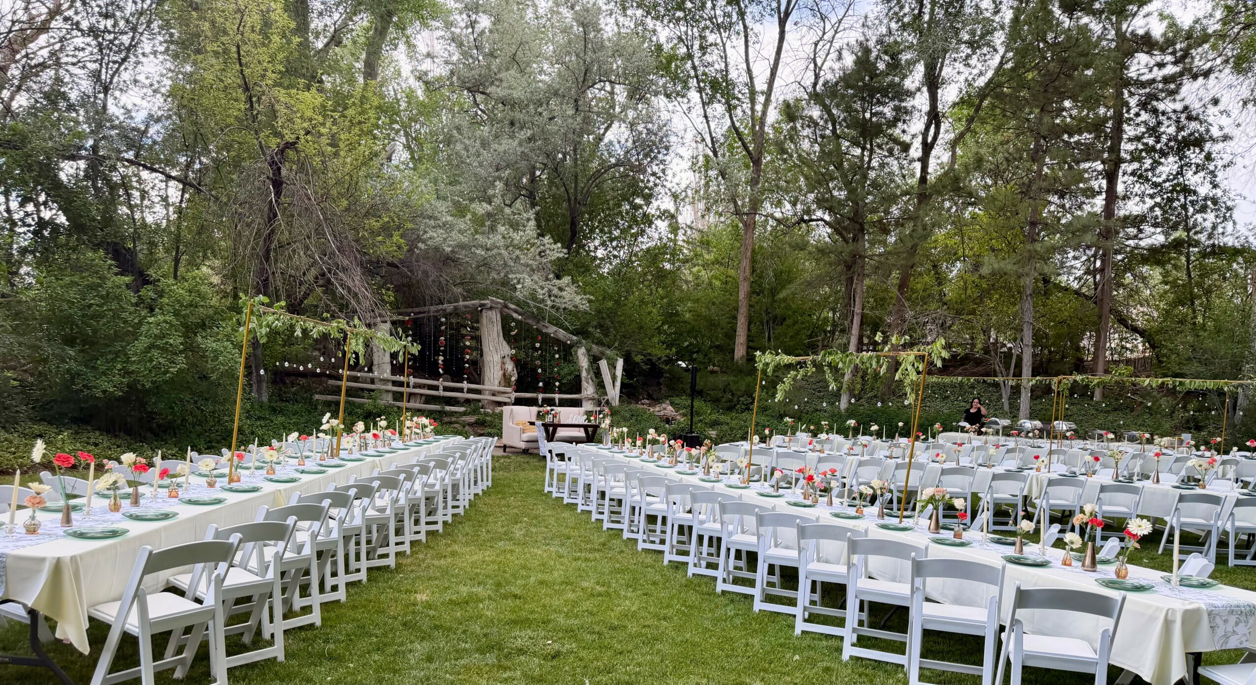 Outdoor event setup with long tables and chairs on grass, surrounded by trees and string lights.