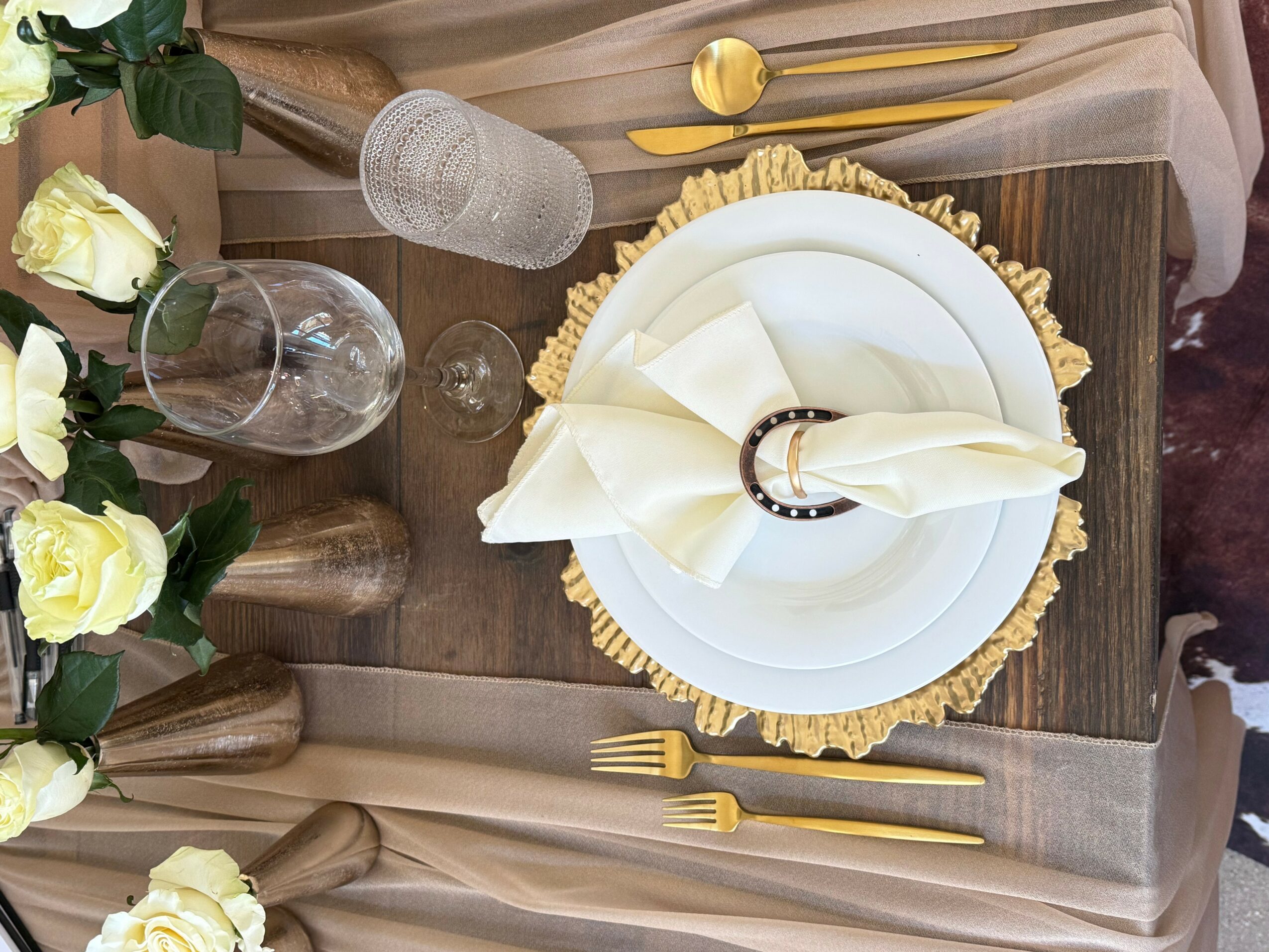 Table setting with white plate, folded napkin, gold cutlery, and decorative placemat, with flowers and glassware nearby.