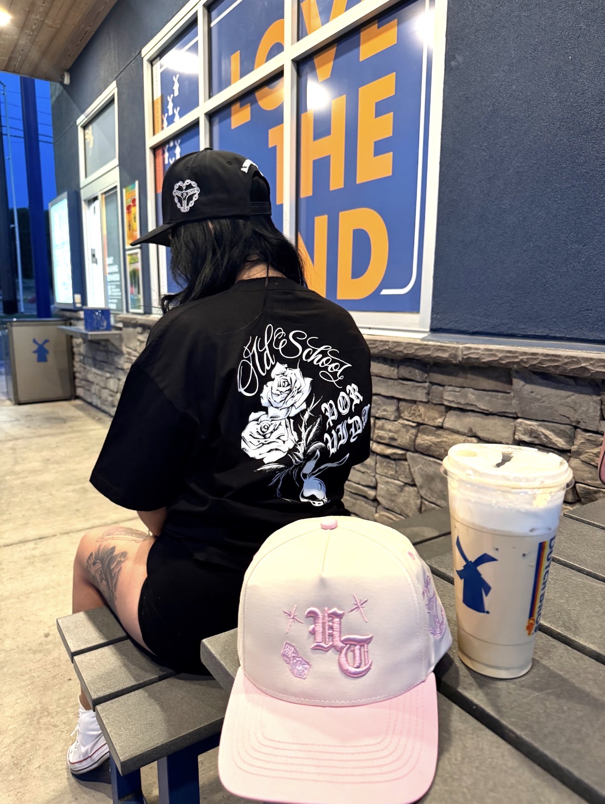 Person sitting at a table with a white cap, black shirt, and a tattooed arm, in an indoor setting with a large drink.