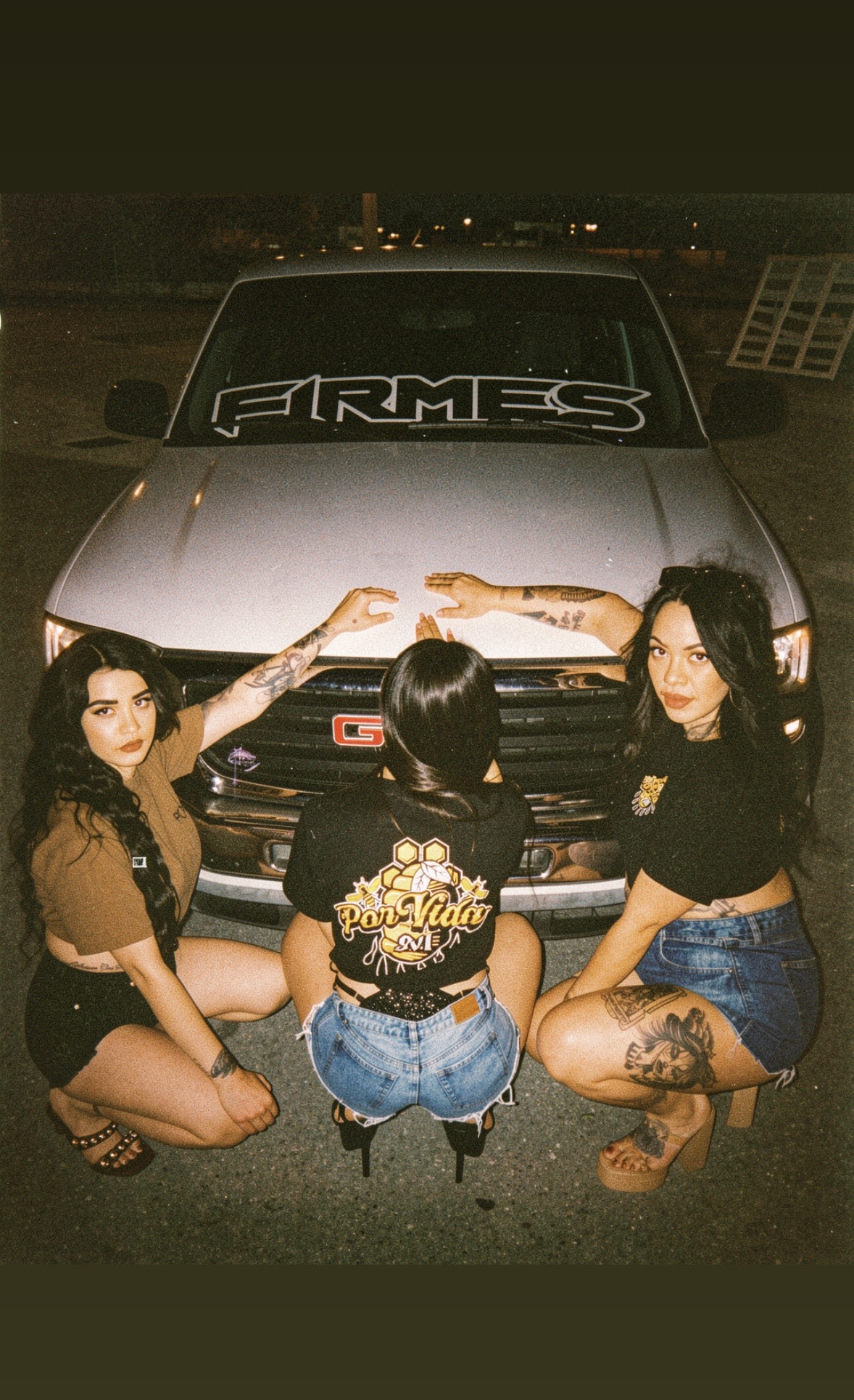 Three women pose in front of a car at night, with city lights in the background. One woman has tattoos and is pointing at the car's windshield. The other two are sitting on the ground, facing the camera.