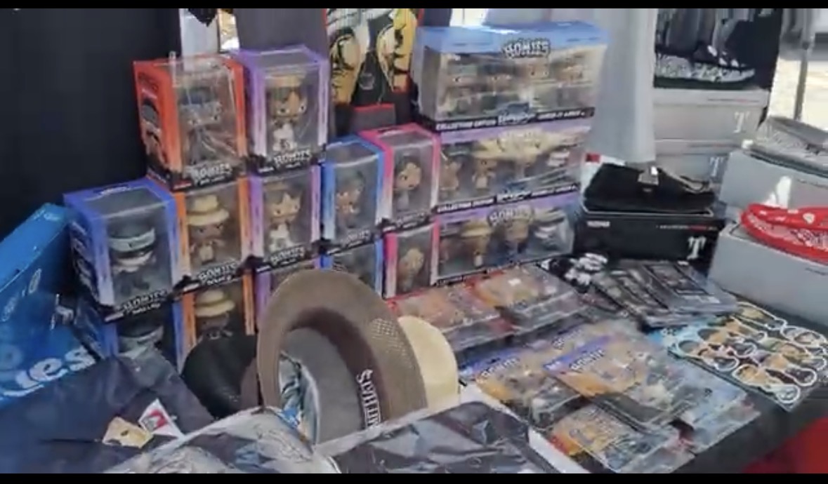 Table with various toys, collectibles, and merchandise displayed, including boxed figures, accessories, and other items.