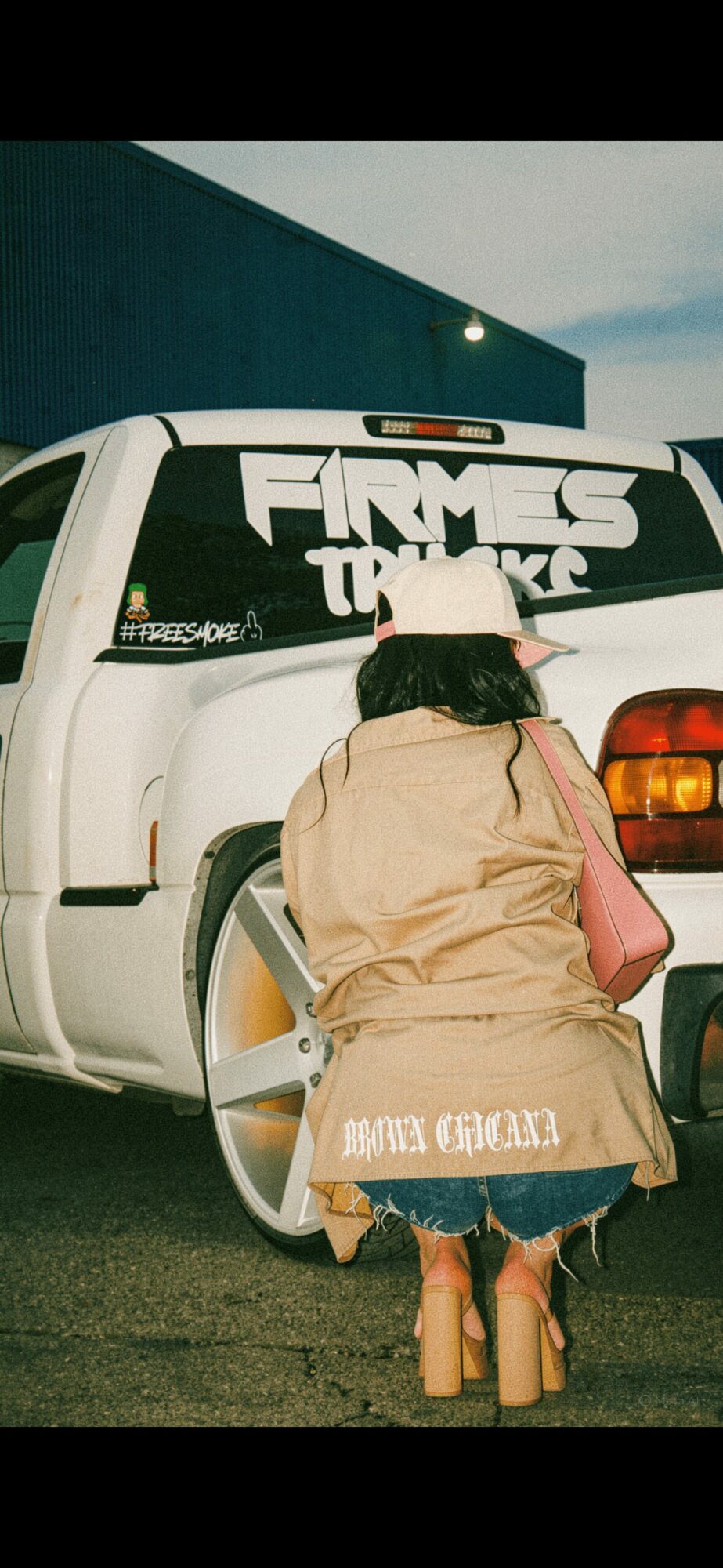 Person with long hair, beige coat, and pink bag stands near a white vehicle with large text on the window, outdoors.