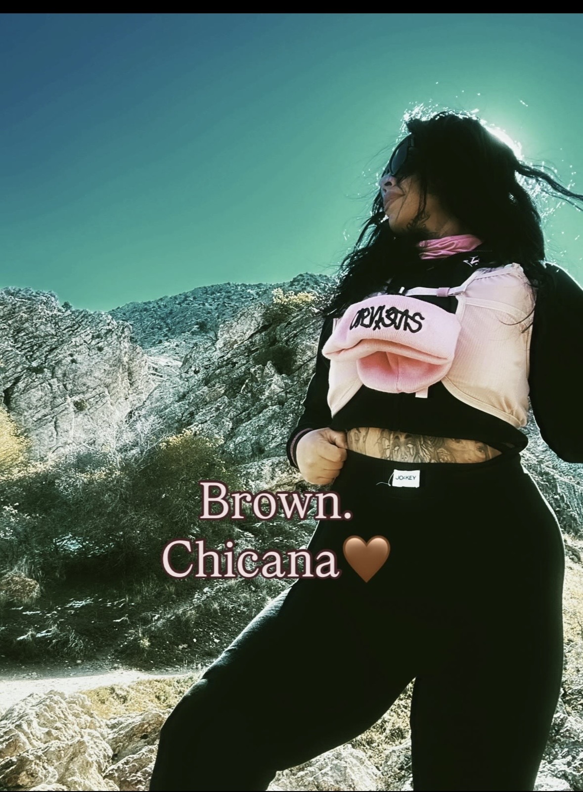 Person with long dark hair standing outdoors near rocks, facing away, wearing a pink and black jacket and black pants.