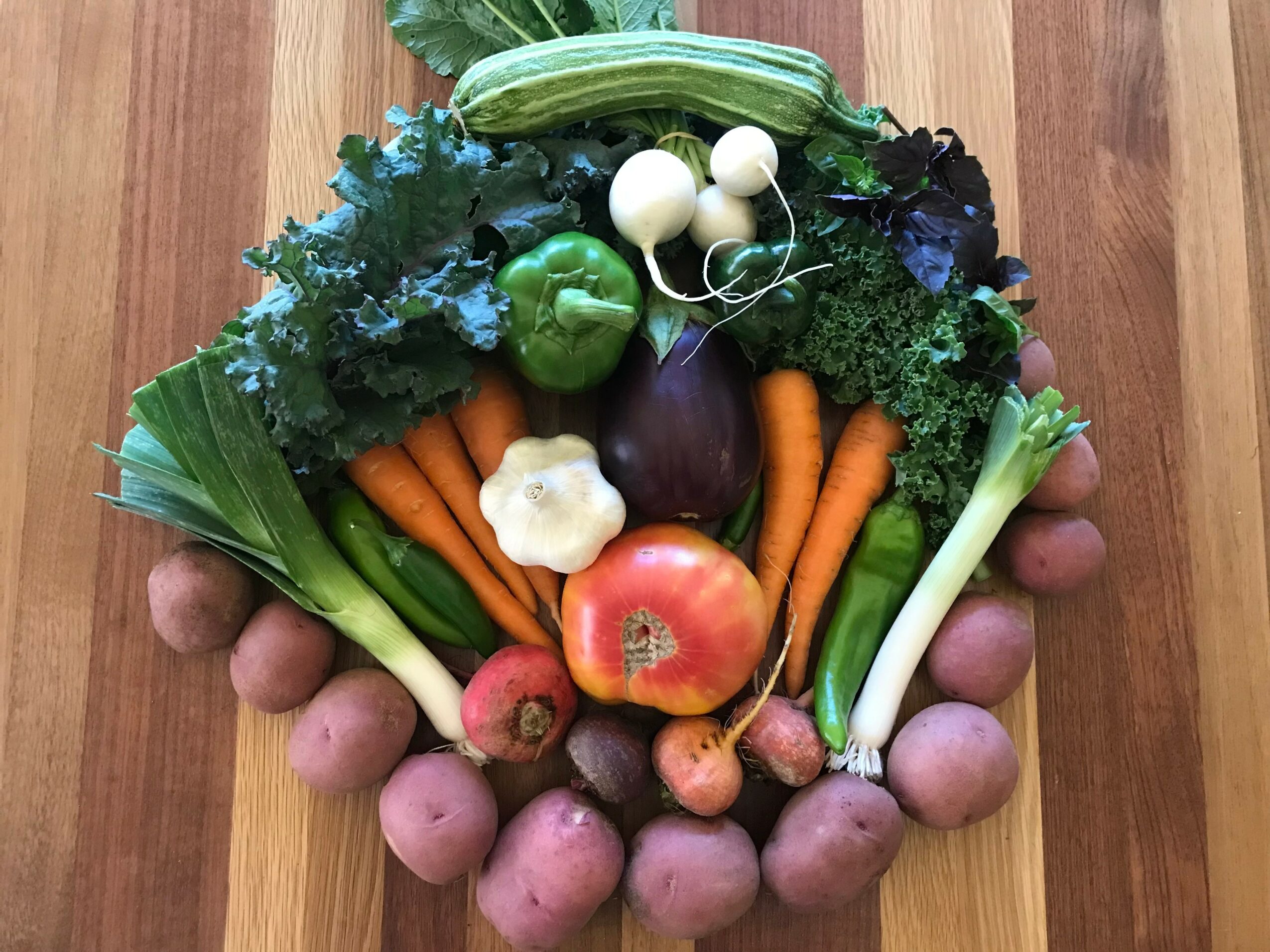 Arrangement of various vegetables including carrots, potatoes, green peppers, onions, garlic, and leafy greens on a wooden surface.