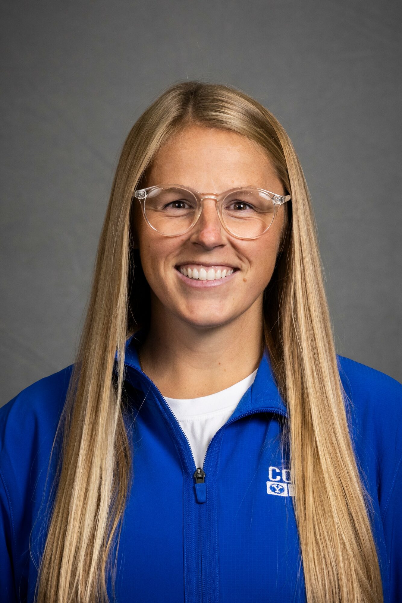 Young woman with long blonde hair wearing glasses and a blue jacket, smiling, against a gray background.