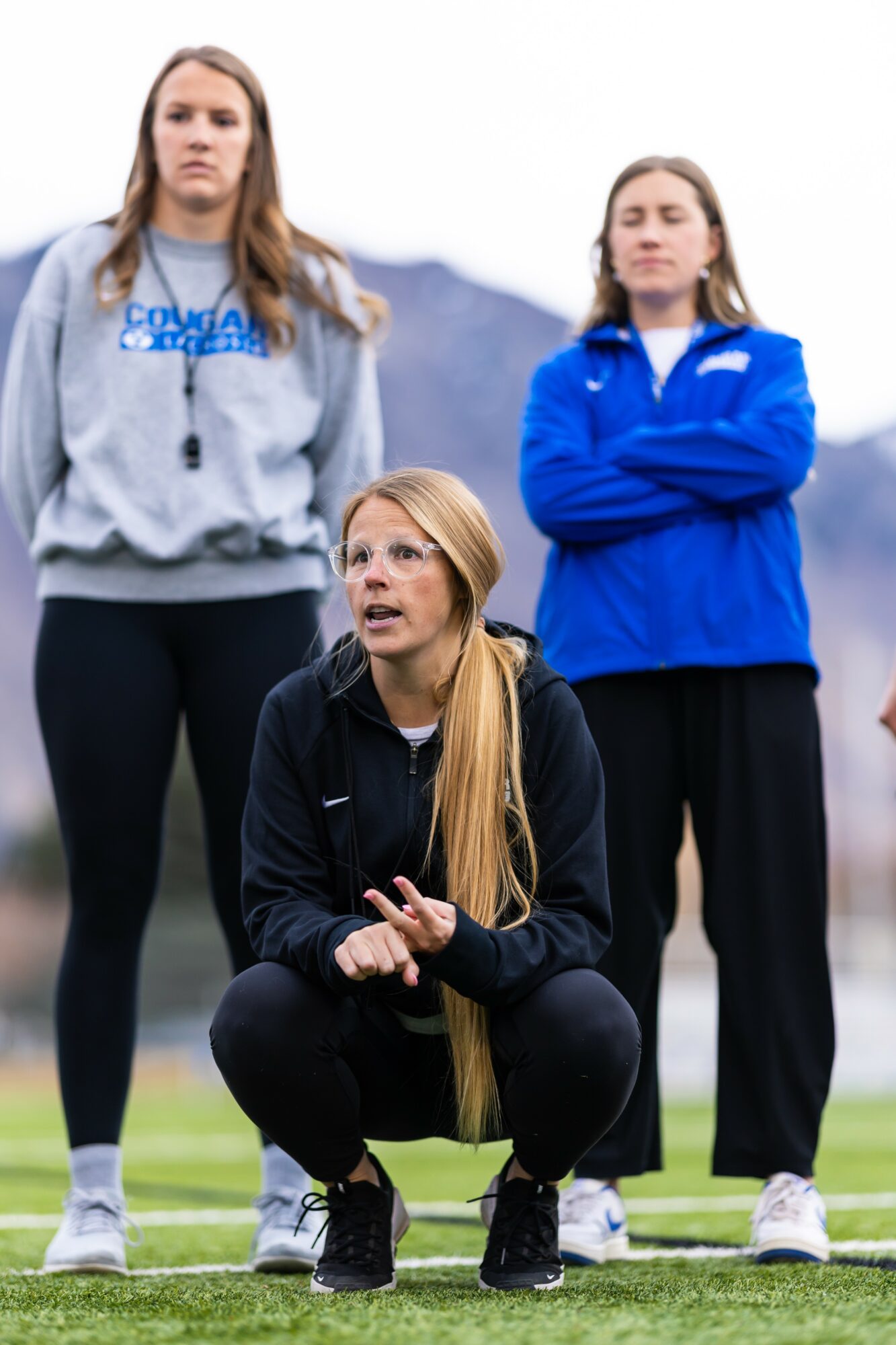 Three women outdoors, one crouching and two standing with arms crossed, mountains in background, cloudy sky.