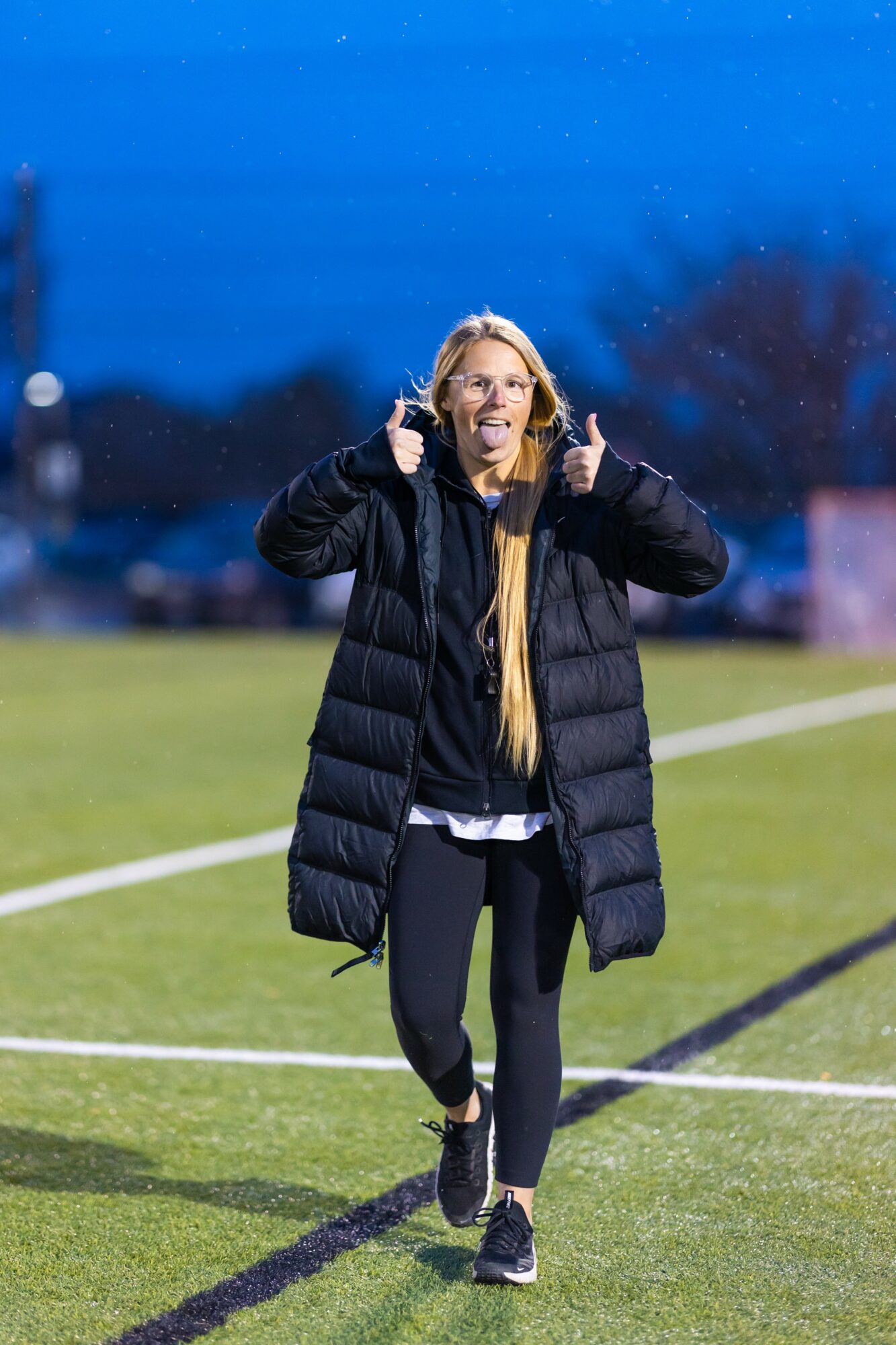 Woman with long blonde hair wearing a black puffer coat, walking on a sports field during evening, smiling, holding hood strings.