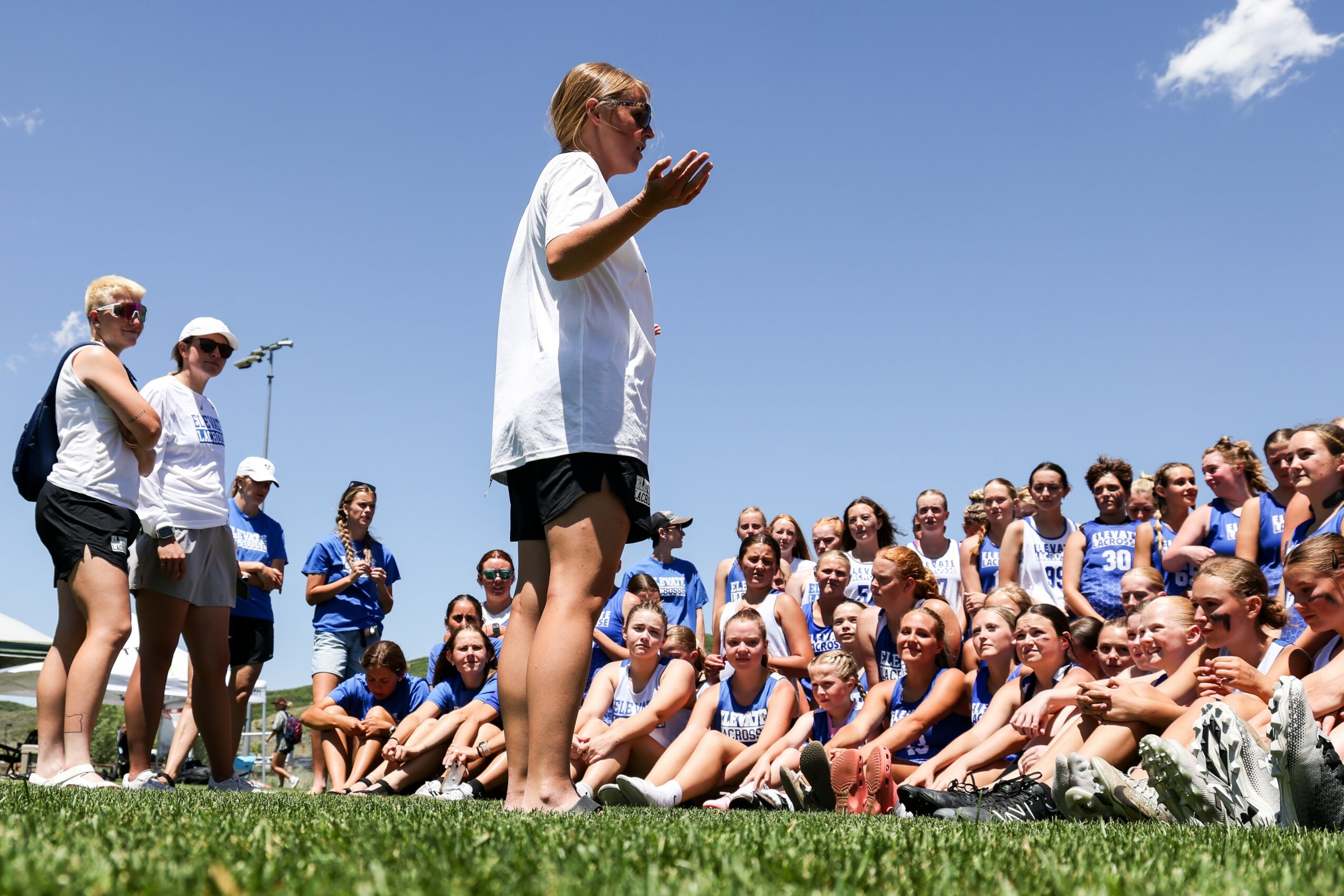 Group of people outdoors on grass, listening to a woman speaking, with a clear blue sky background.