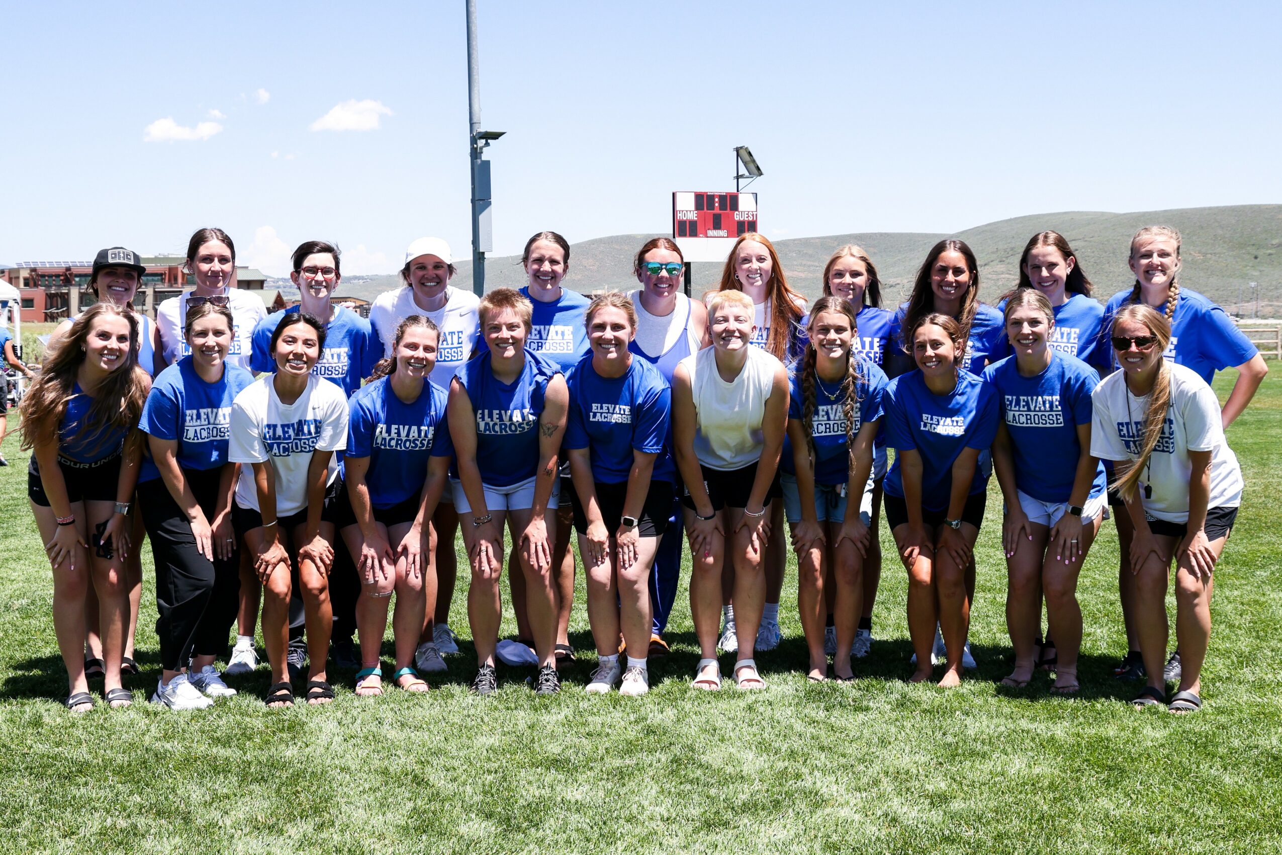 Group of young women and girls in blue sports uniforms standing on a grassy field, smiling, with a clear sky and sports field in background.