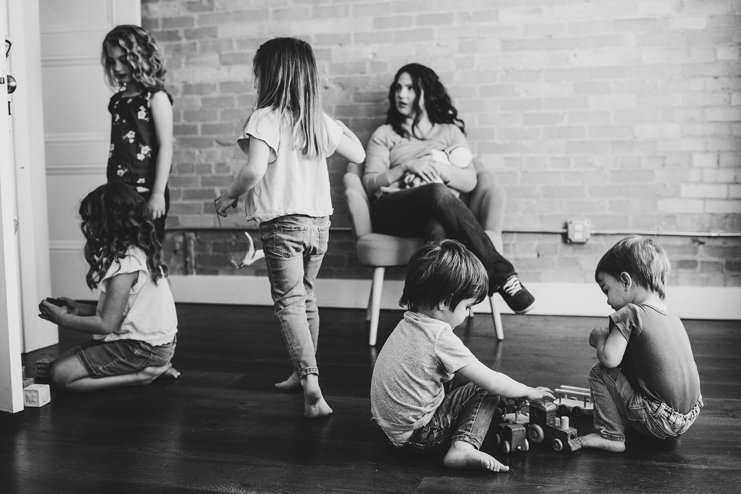 Four children playing on the floor and a woman sitting on a chair against a brick wall, in a room with wooden floor.