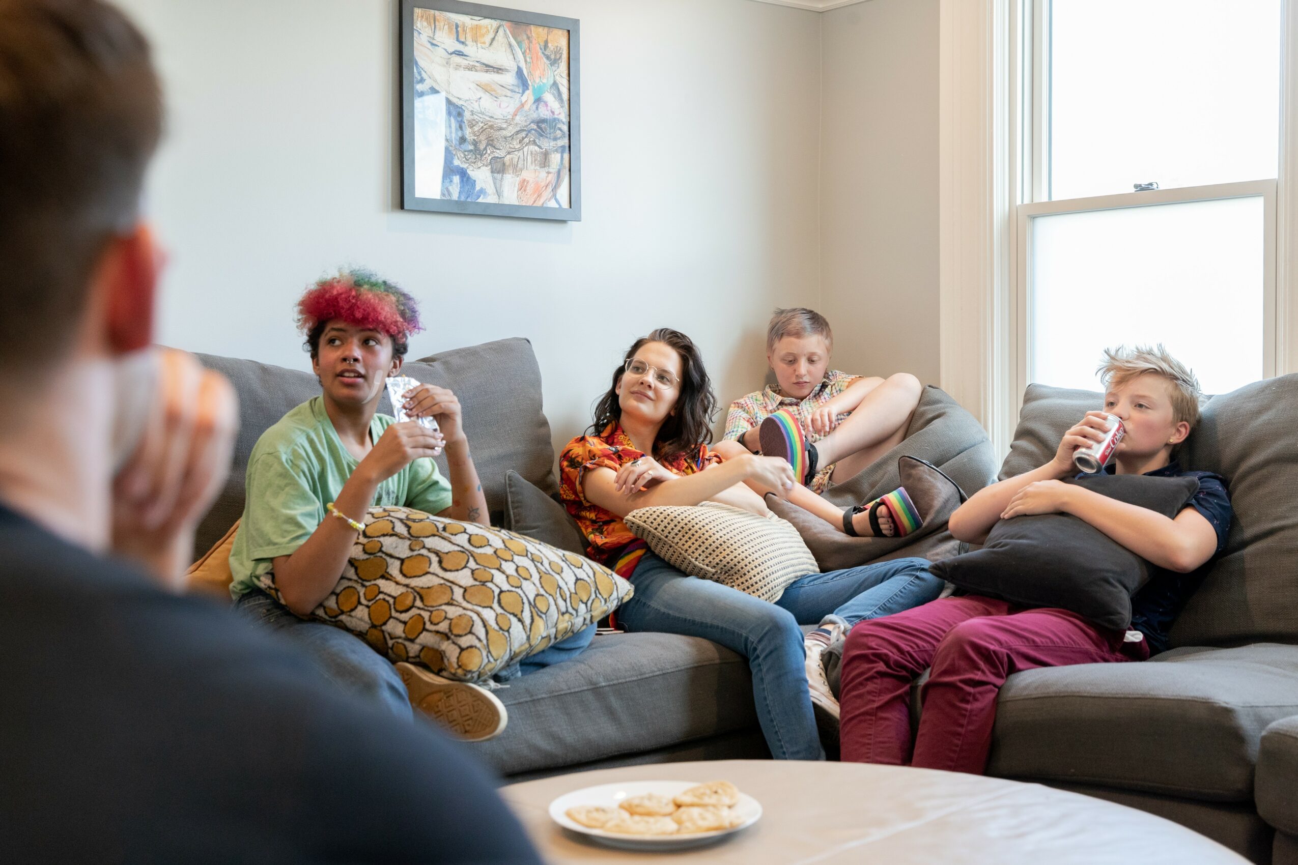 Group of five people sitting on a gray couch in a living room, talking and relaxing, with a window and artwork on the wall.