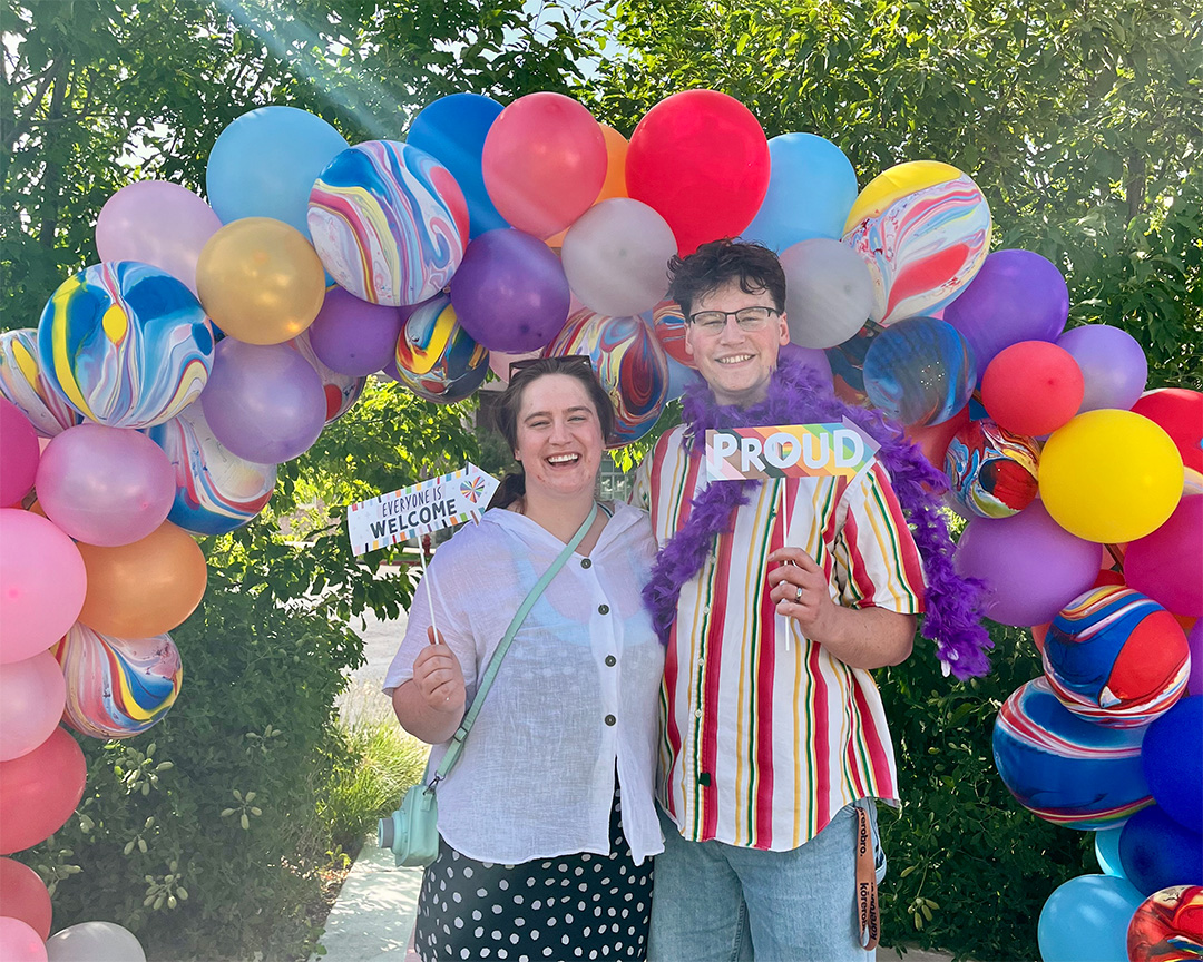 Two people stand under a colorful balloon arch outdoors, smiling, with trees and sunlight in the background.