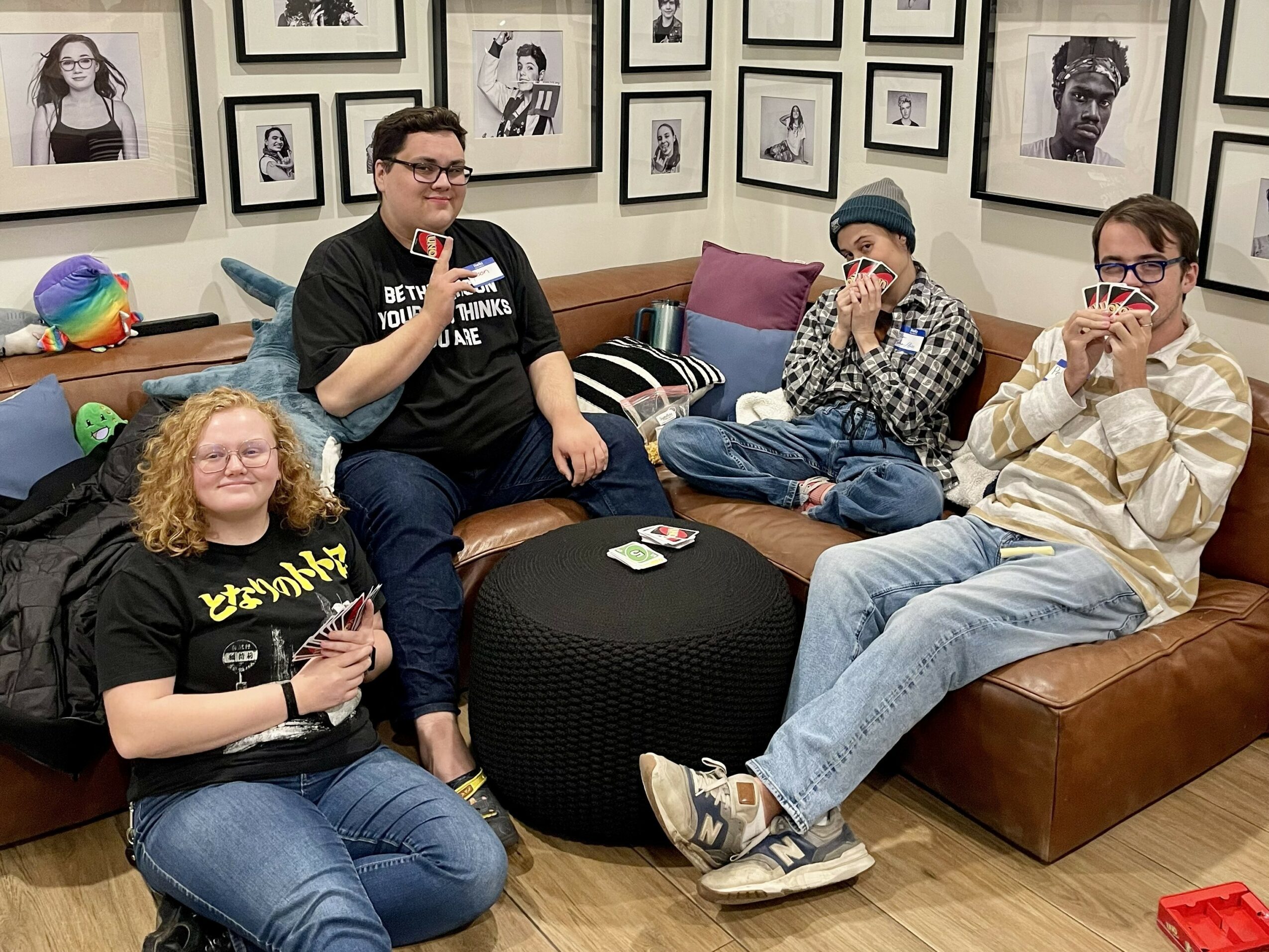 Four people sitting on couches in a room with framed photos on the wall, some holding cups or phones.