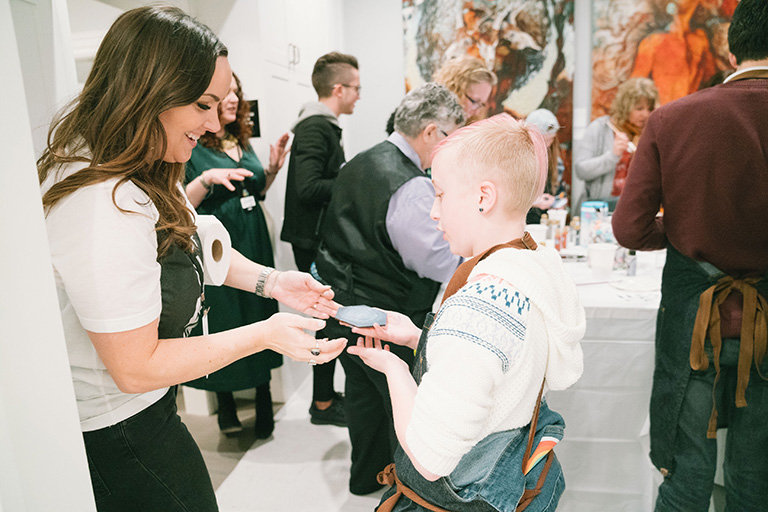 Woman handing a small object to a young boy in a crowded art gallery with colorful paintings, people in the background.