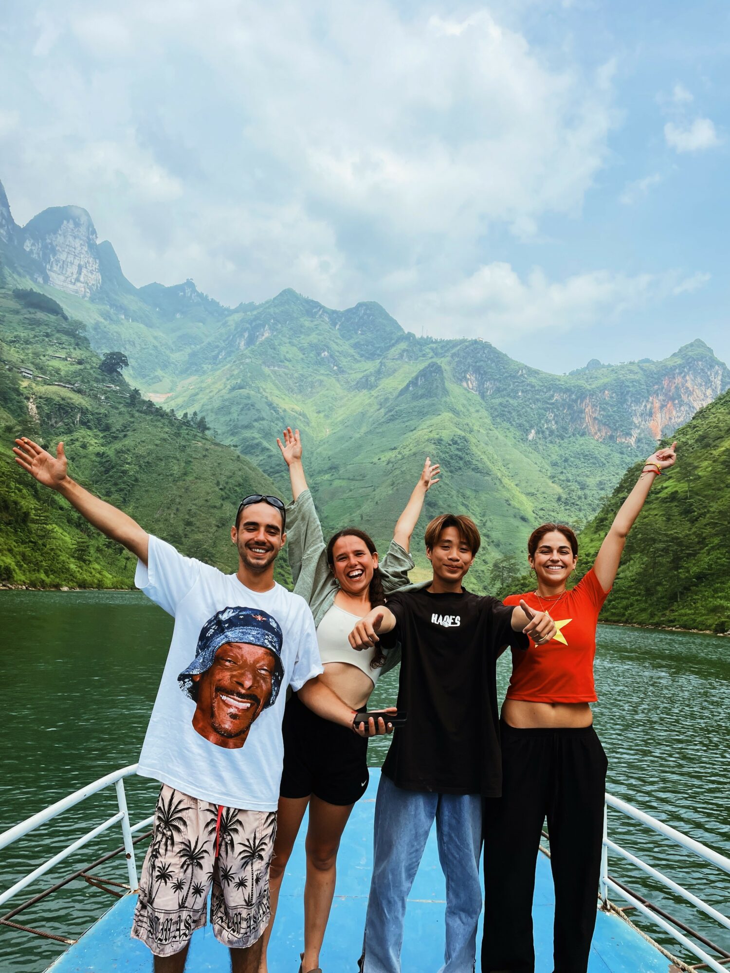 Four people on a boat with mountains and a river in the background, smiling and raising their arms.