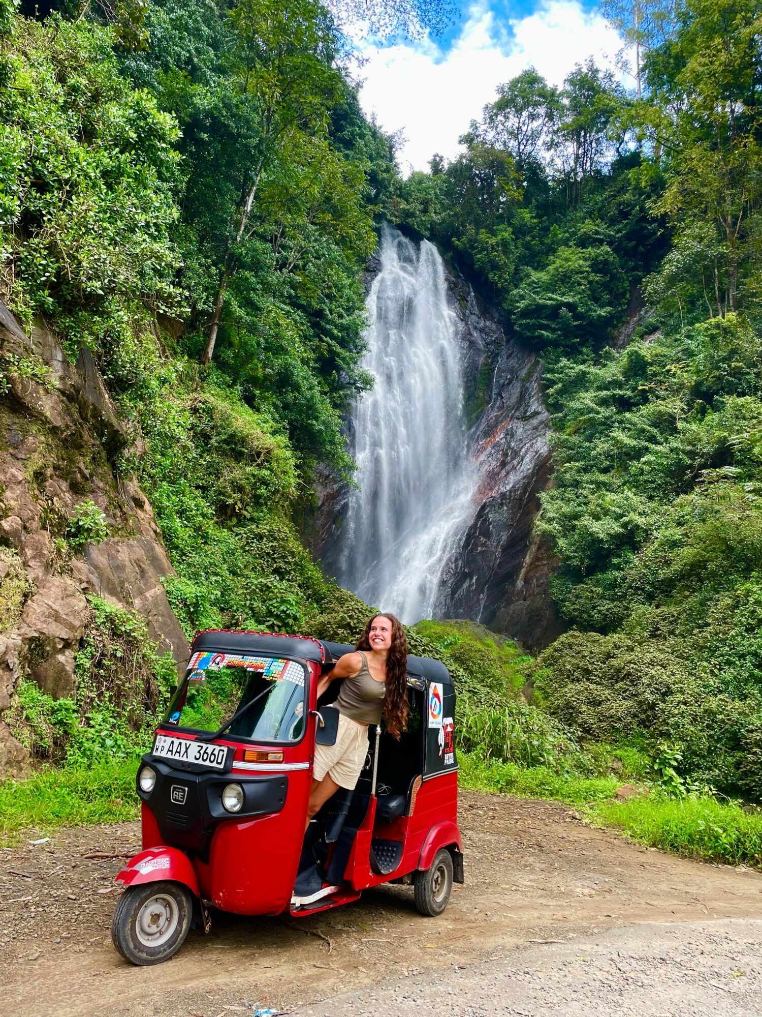 Person standing next to a red auto rickshaw in front of a waterfall surrounded by green trees.