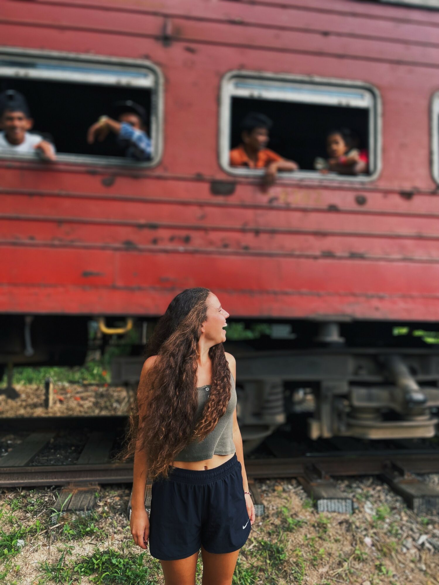 A woman with long, curly hair stands near train tracks, looking at children in a red train carriage window.