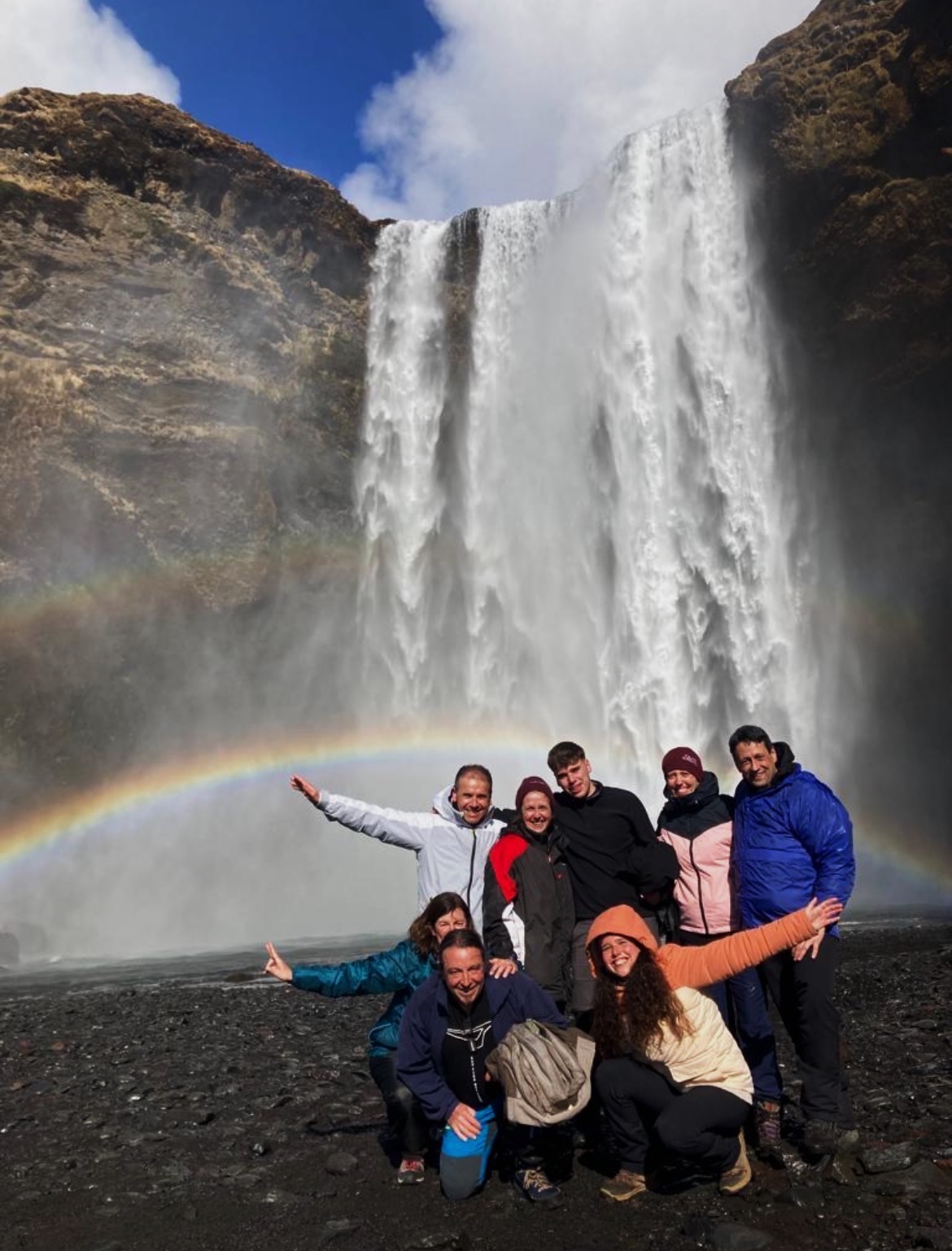 Group of people posing in front of a large waterfall with a rainbow, some with arms outstretched, on dark ground.