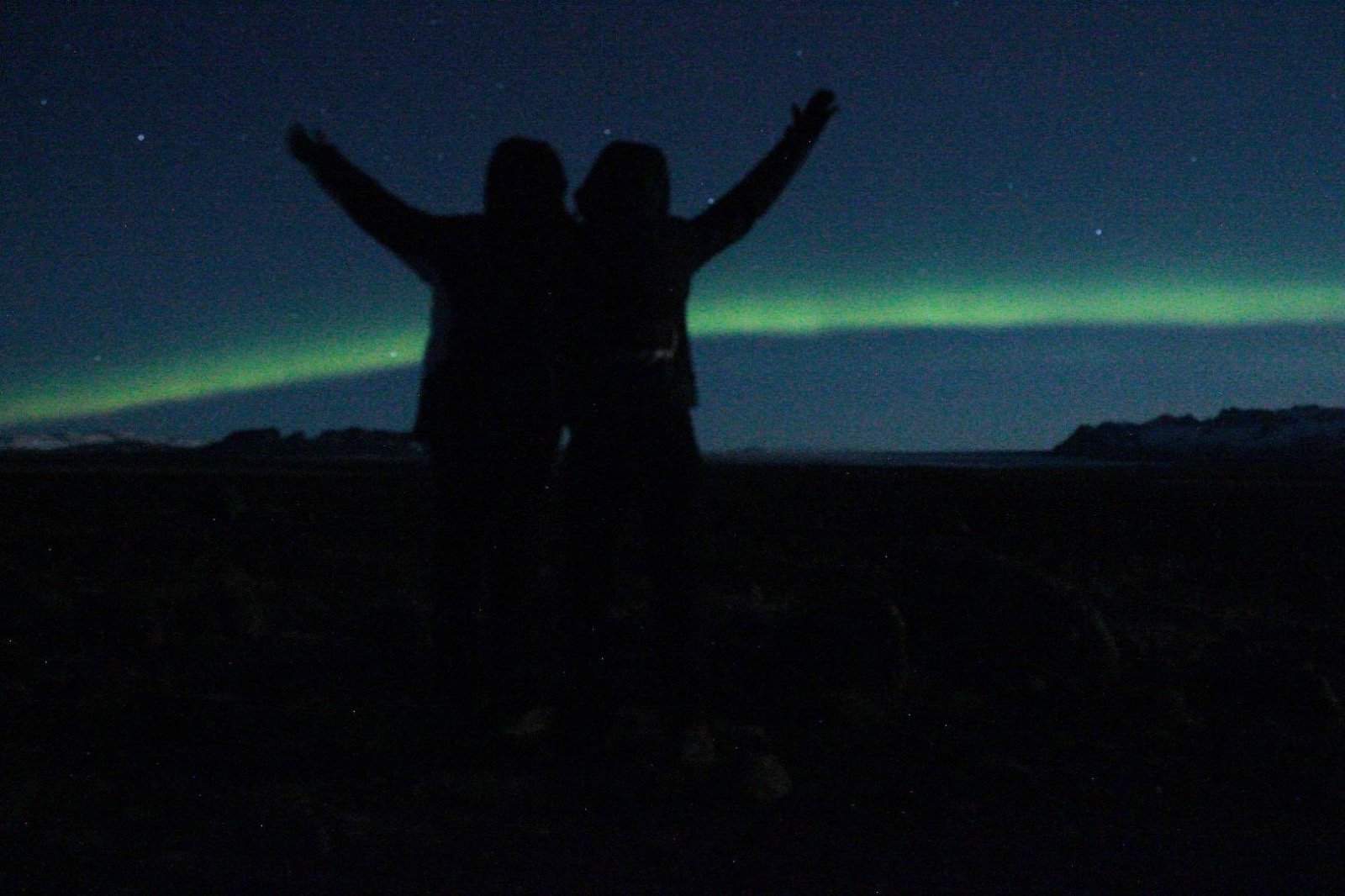 Two people stand outdoors at night with arms raised, northern lights visible in the sky.