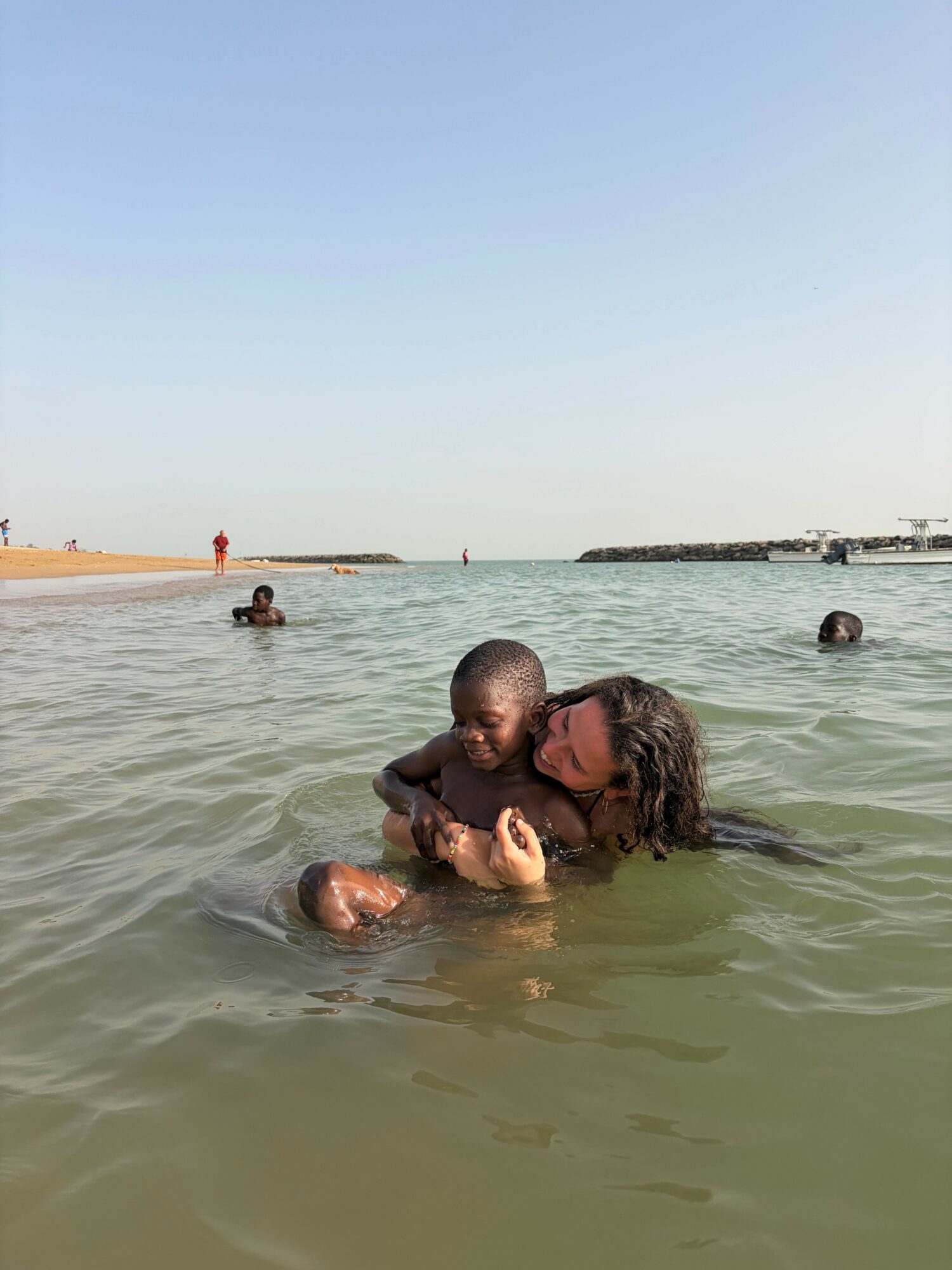 Two people embrace in water, with others in the background, under a clear sky.
