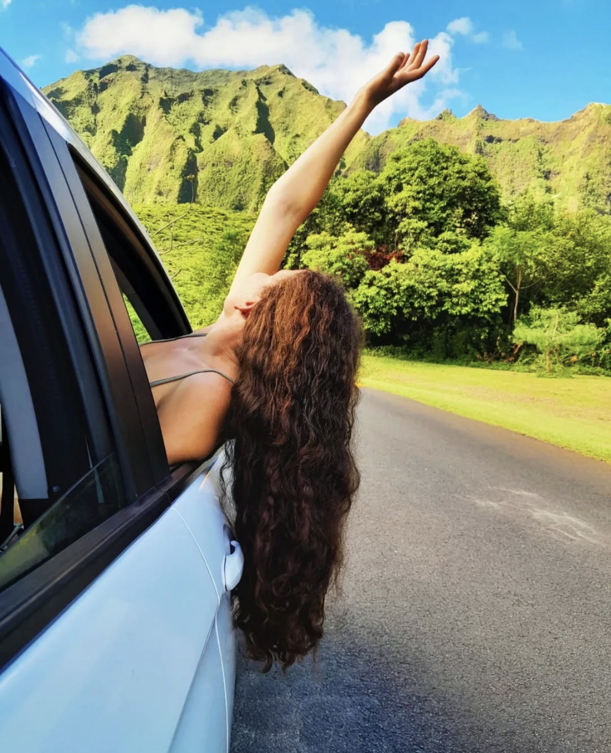 Person with long hair leaning out of car window, arm raised, scenic green mountains and trees in background.