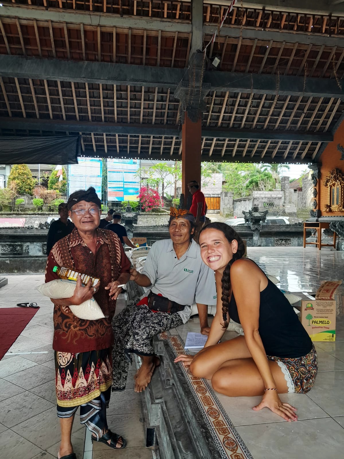 Three women sitting on a patterned rug under a wooden roof, smiling, with a water feature and trees in the background.