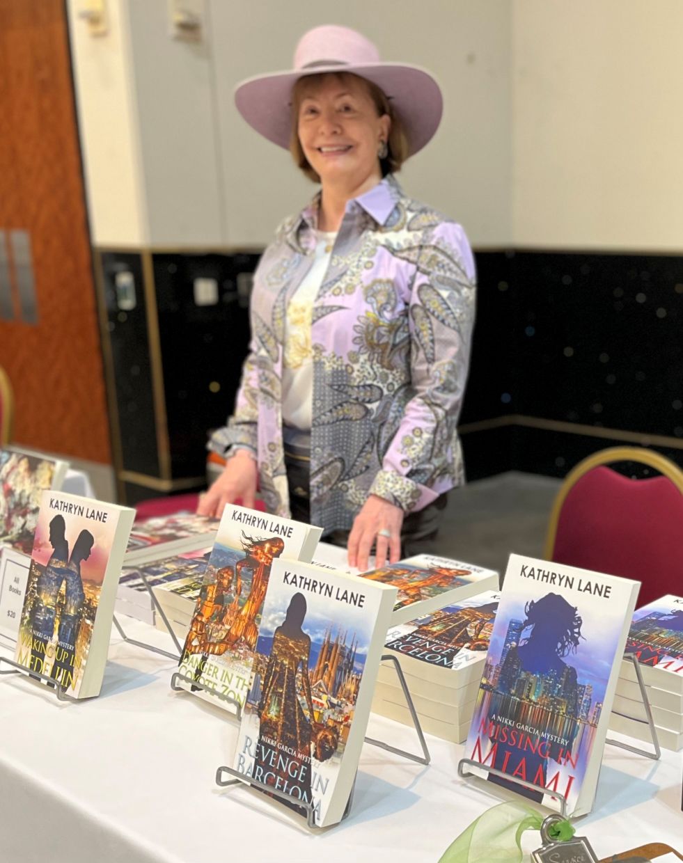 Smiling woman wearing a large pink hat and patterned jacket standing behind a table with books displayed.