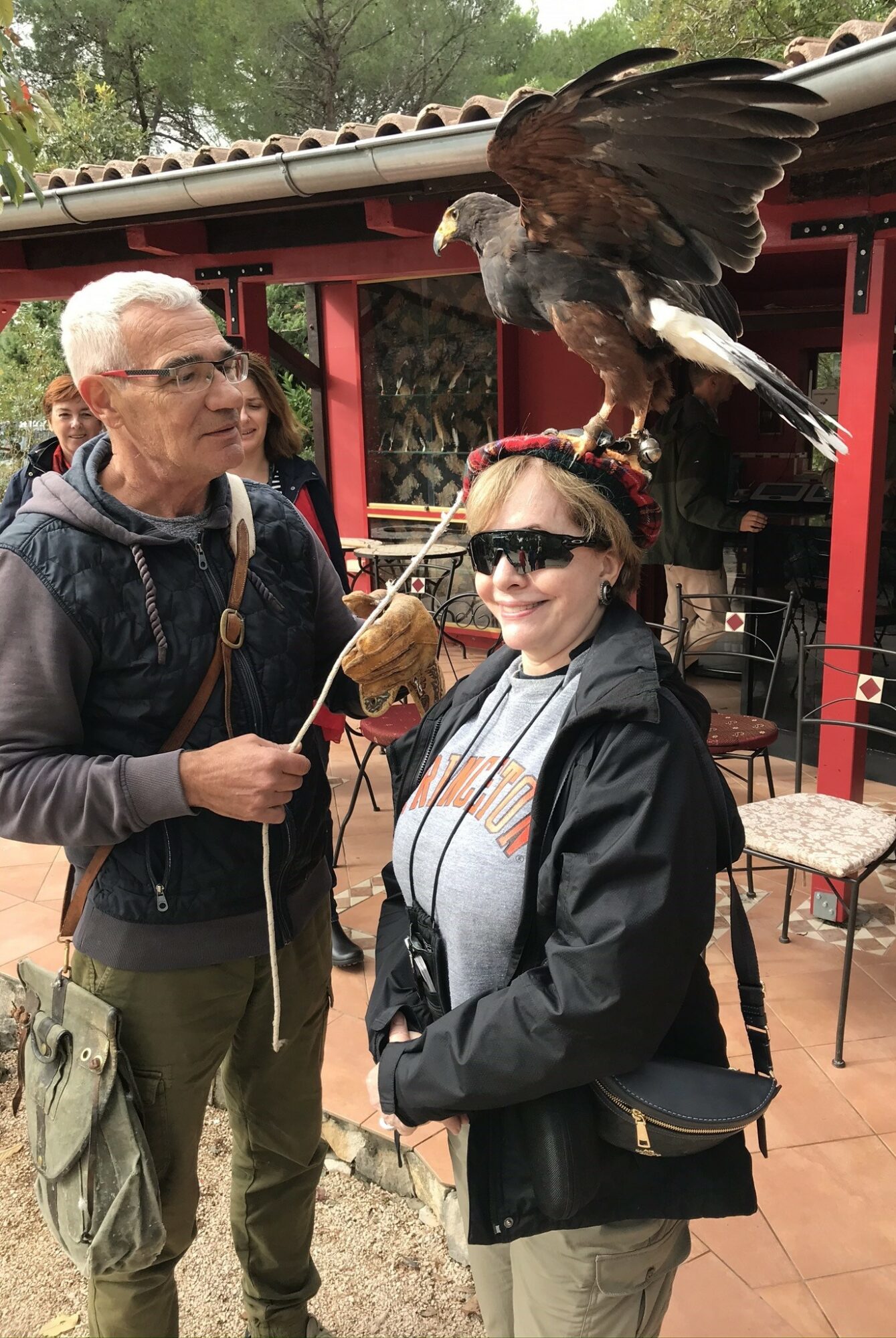 Two people, one with sunglasses, stand outdoors with a large bird perched on a person's arm, in front of a red structure.