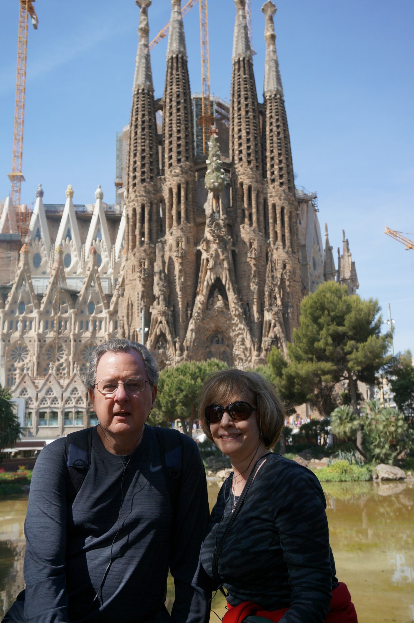 Two people standing in front of Sagrada Família basilica with trees and a pond, smiling at the camera.