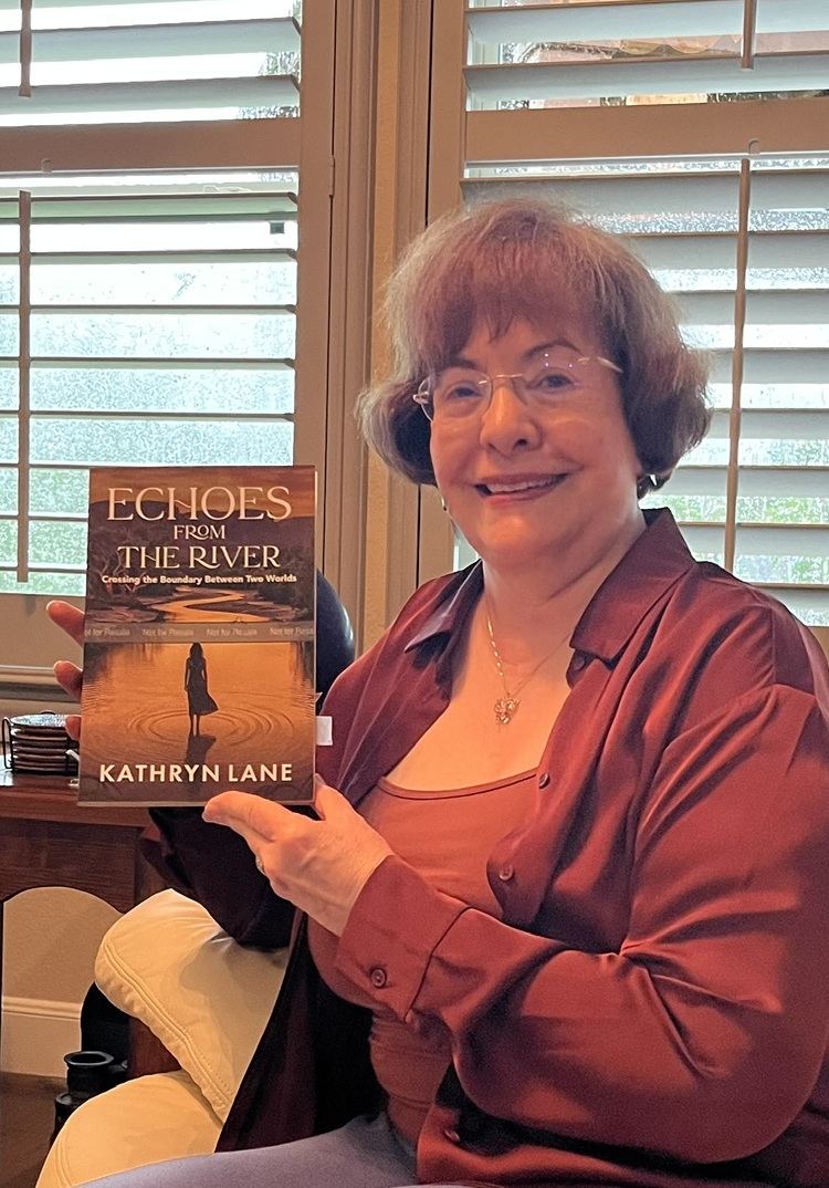 Older woman with glasses and short hair holding a book titled 'ECHOES FROM THE RIVER' in front of window blinds.