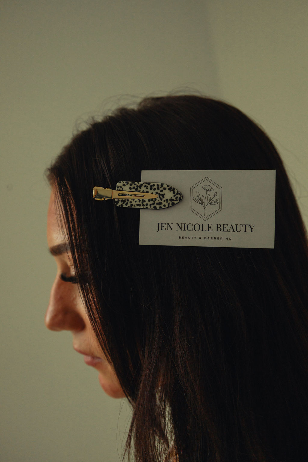 Side profile of a woman with dark hair and a hair clip holding a business card that reads 'Jen Nicole Beauty'.