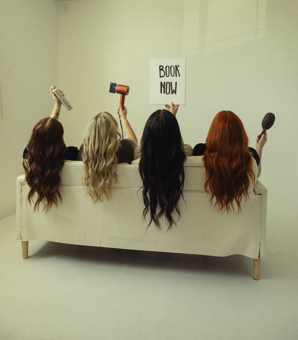 Four women with different hair colors sit on a white couch, holding various items, with a sign reading 'BOOK NOW' behind them.