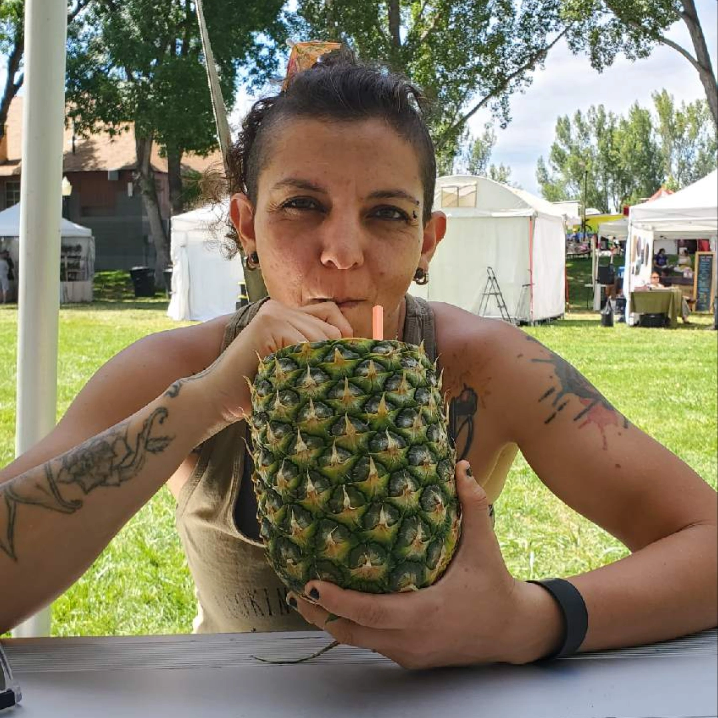 Woman sitting outdoors at a table, holding a pineapple-shaped drink with a straw, surrounded by trees and tents.