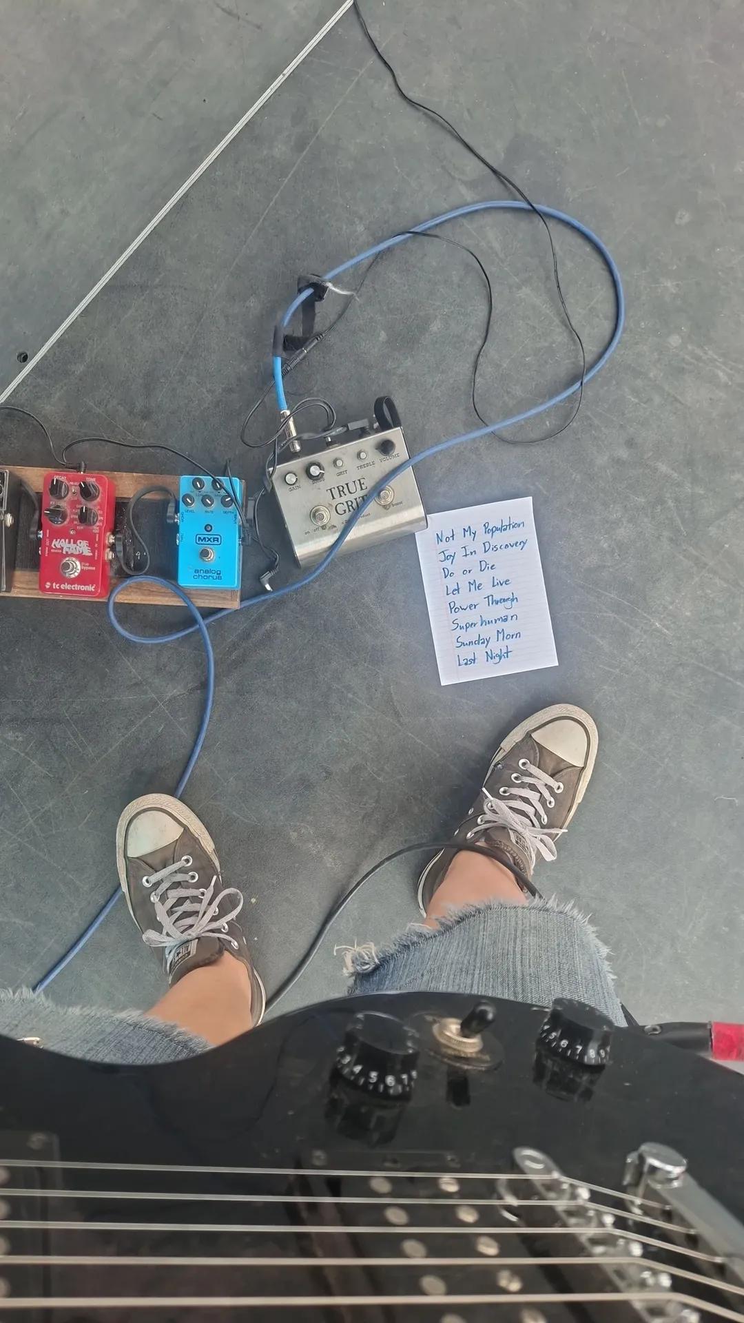 Person wearing sneakers standing on a gray floor with guitar pedals and a handwritten note nearby.
