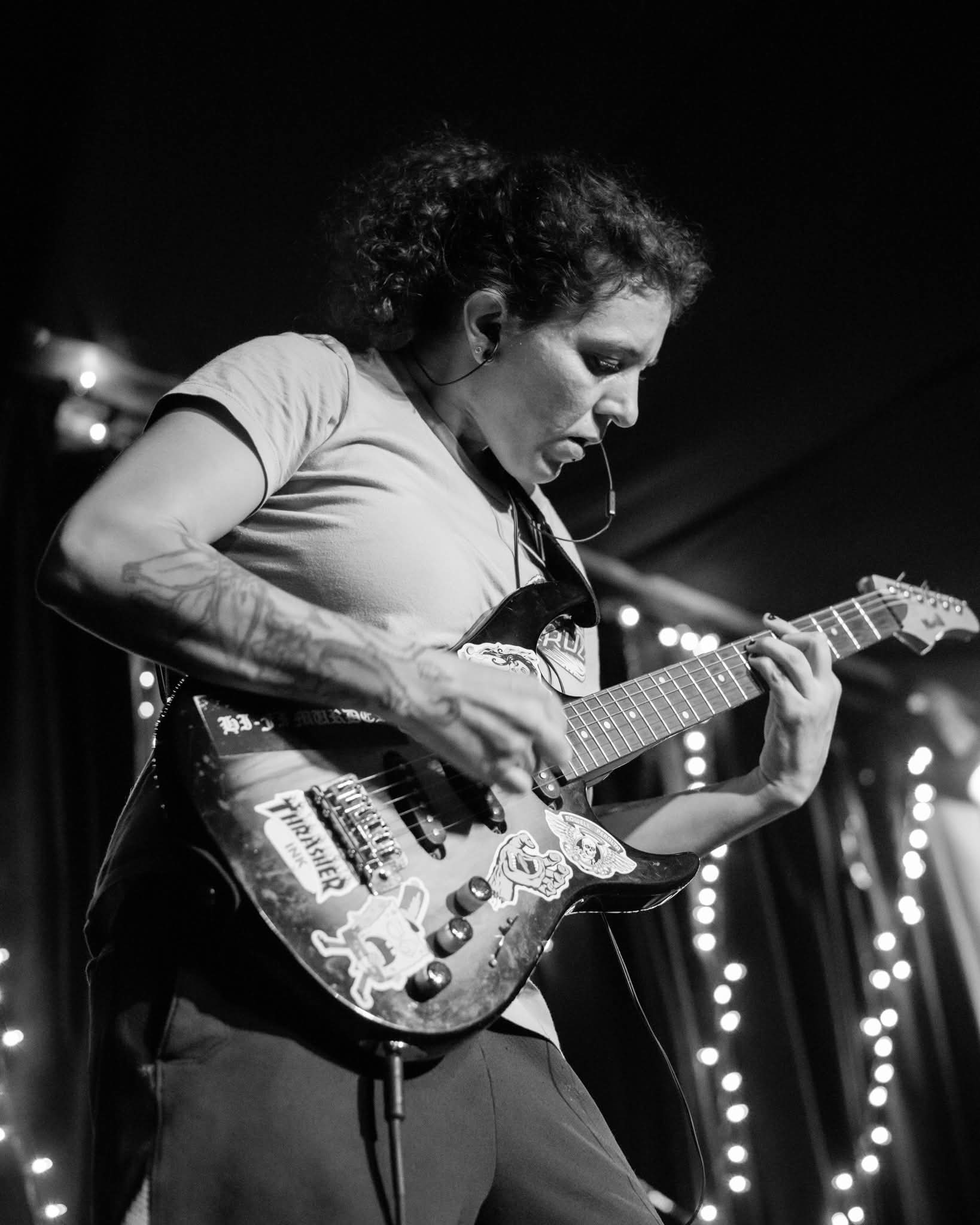 Person playing an electric guitar on stage with lights in background, black and white image.