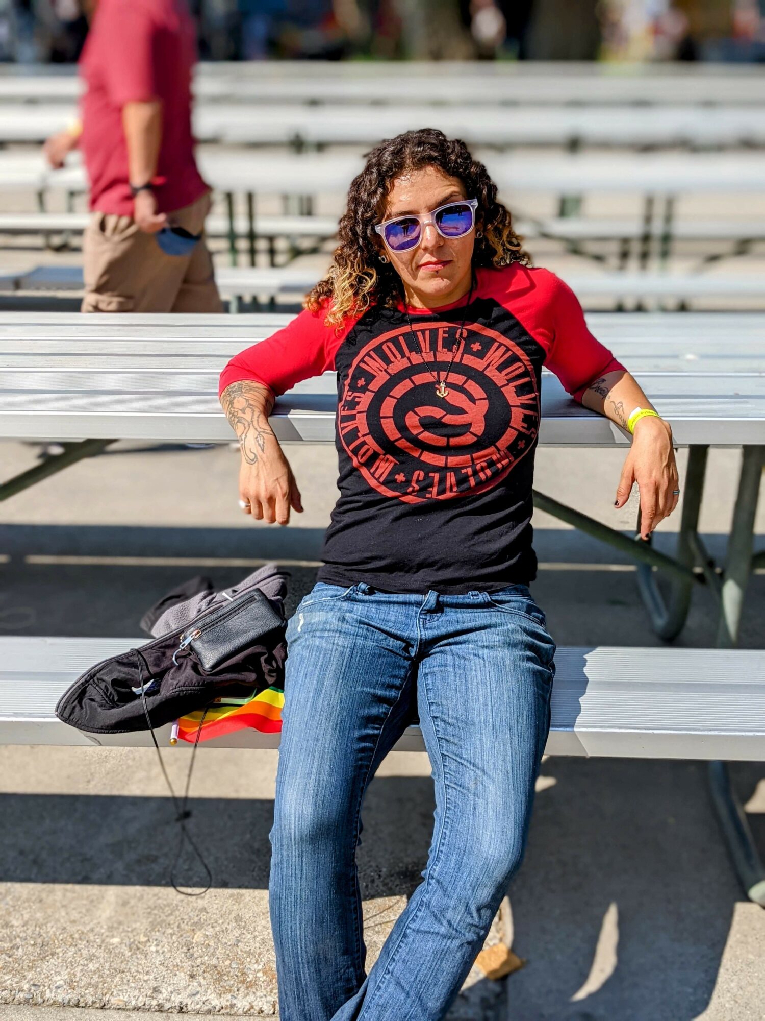 Woman with curly hair wearing sunglasses, red and black shirt, sitting on a bench outdoors.