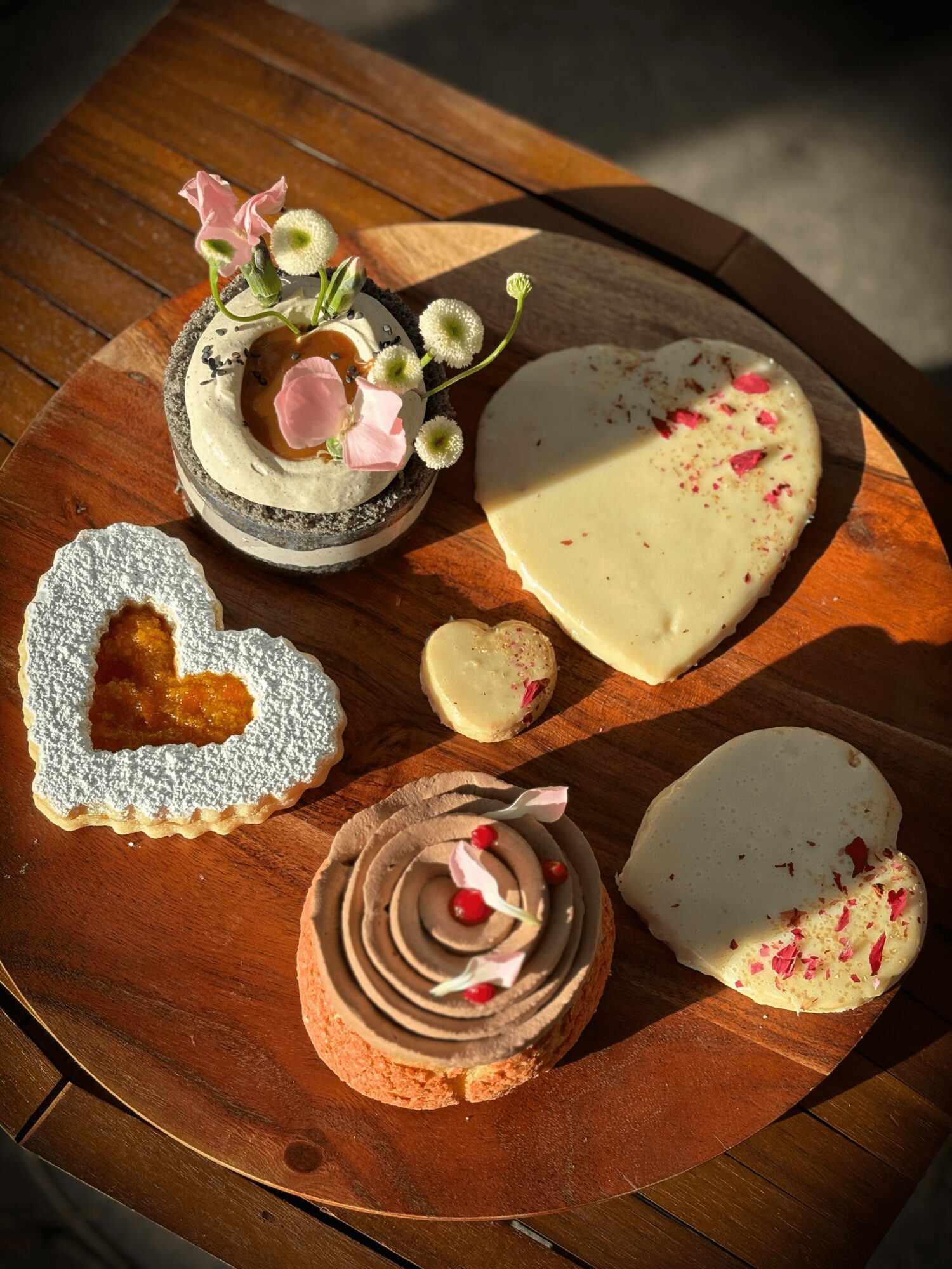 Heart-shaped white chocolate with pink sprinkles, round pink and white cupcake with red heart, heart-shaped lace cookie, small cake with pink flowers, and a flower arrangement on a wooden surface.