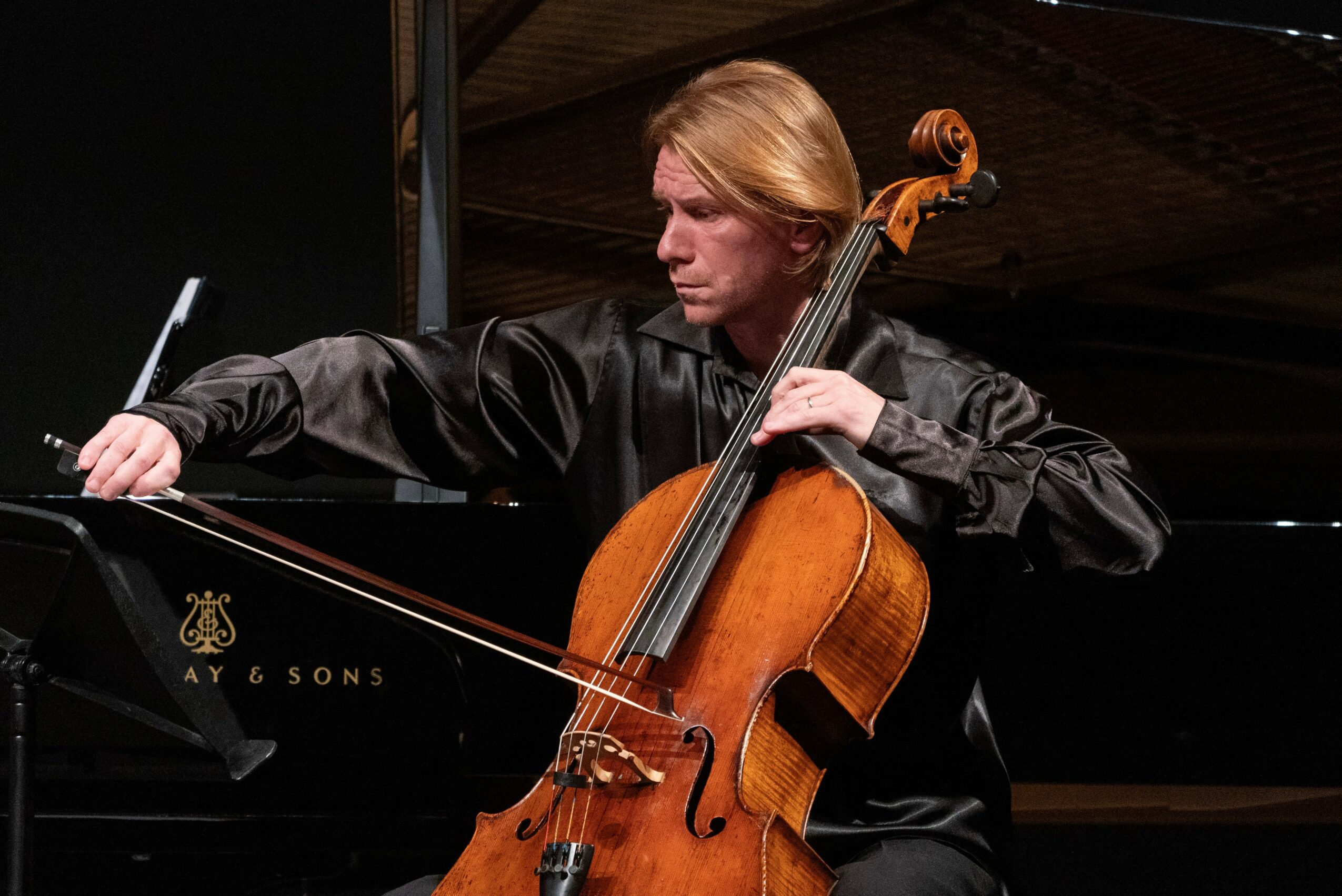 Person playing a cello on stage with a grand piano in the background, wearing a black outfit.
