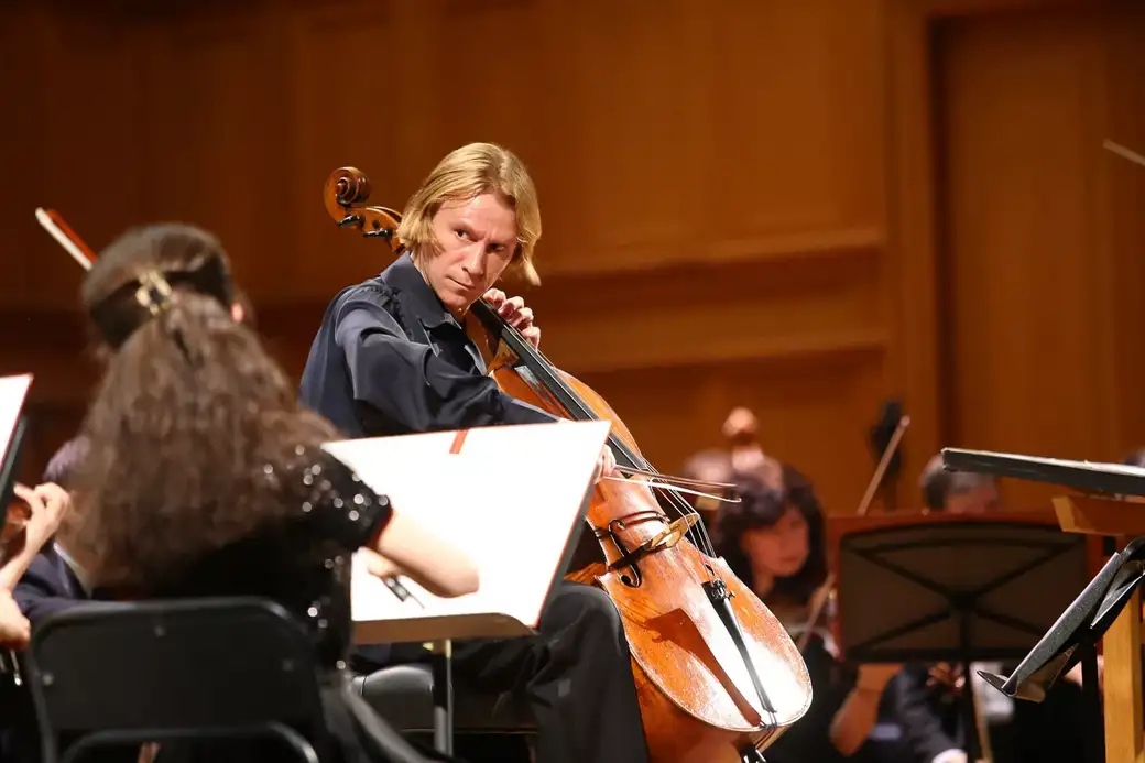 Woman playing cello in a concert hall with other musicians and music stands around her.