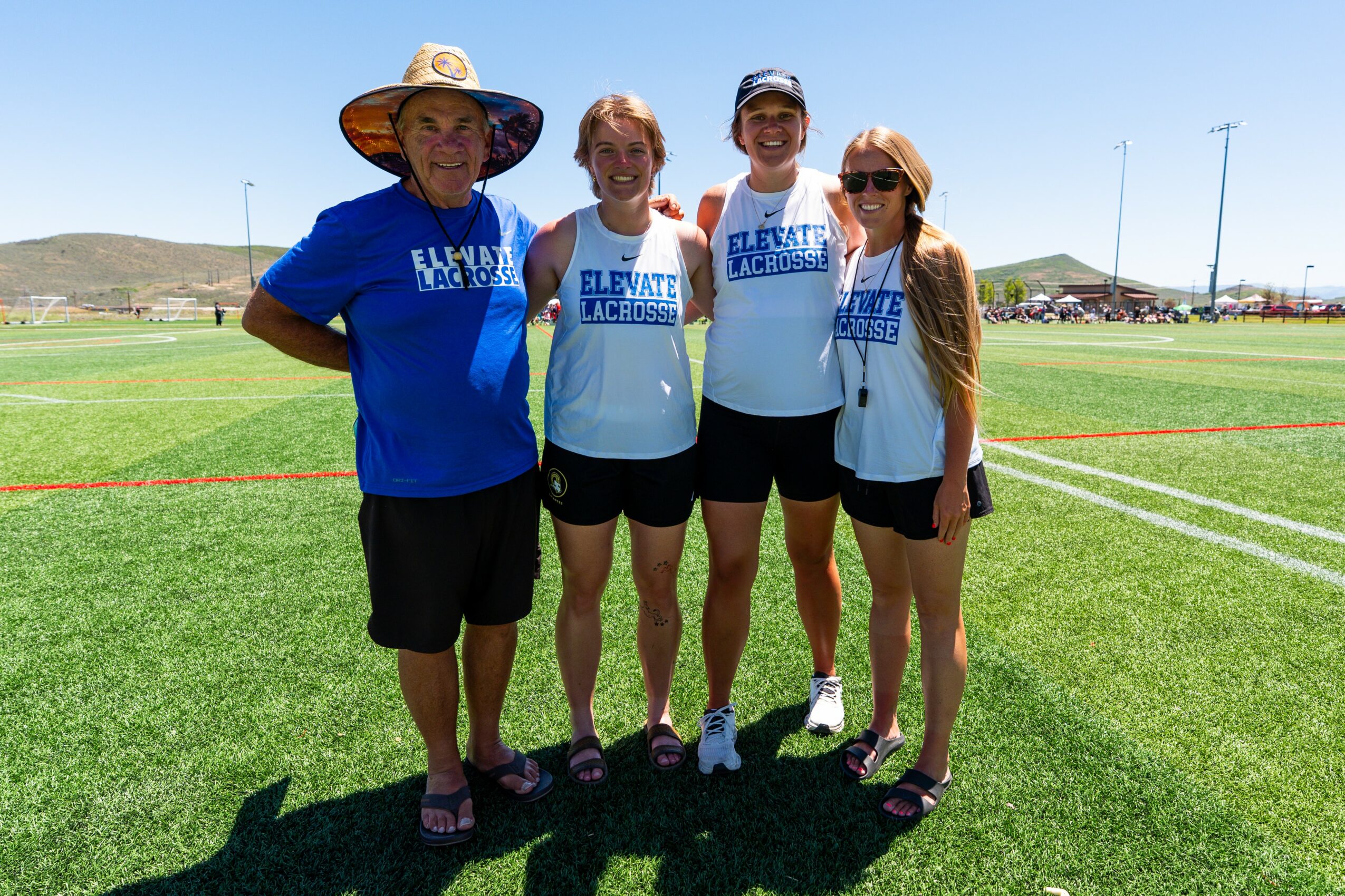 Four people standing on a grassy field, smiling, with a clear sky and sports field in the background.