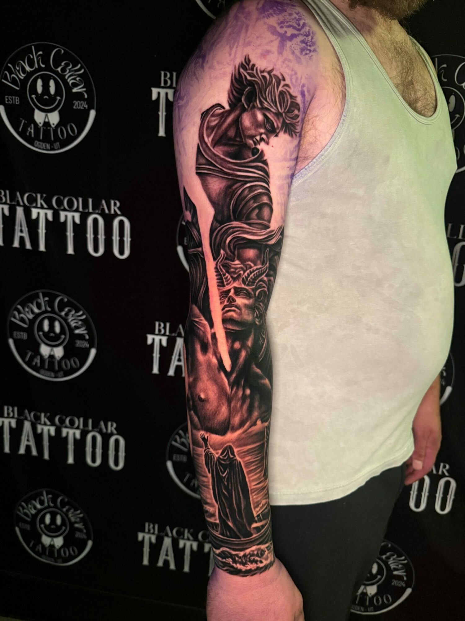 Person lying on a surface with a detailed black and gray tattoo sleeve on their arm, featuring a woman and architectural elements.